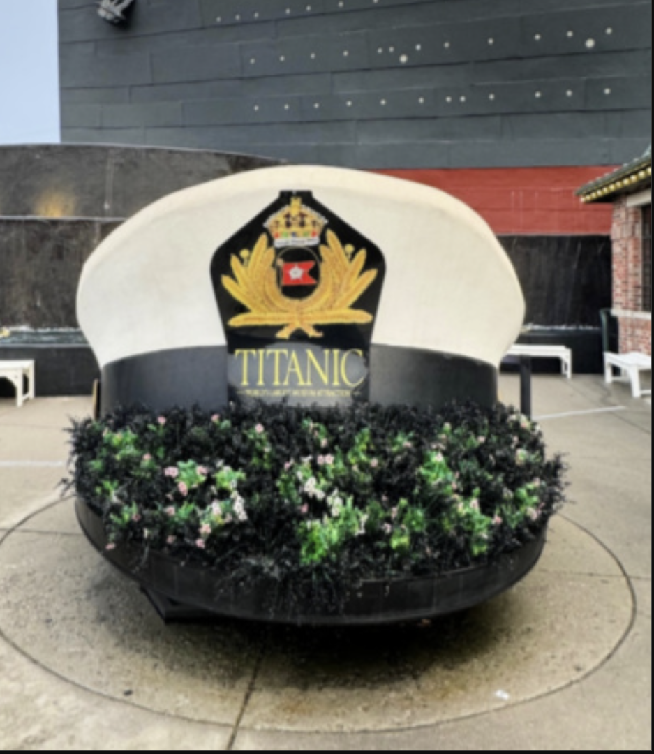 A decorative sign for Titanic, surrounded by flowers, with a crest or emblem above the name, situated outdoors.
