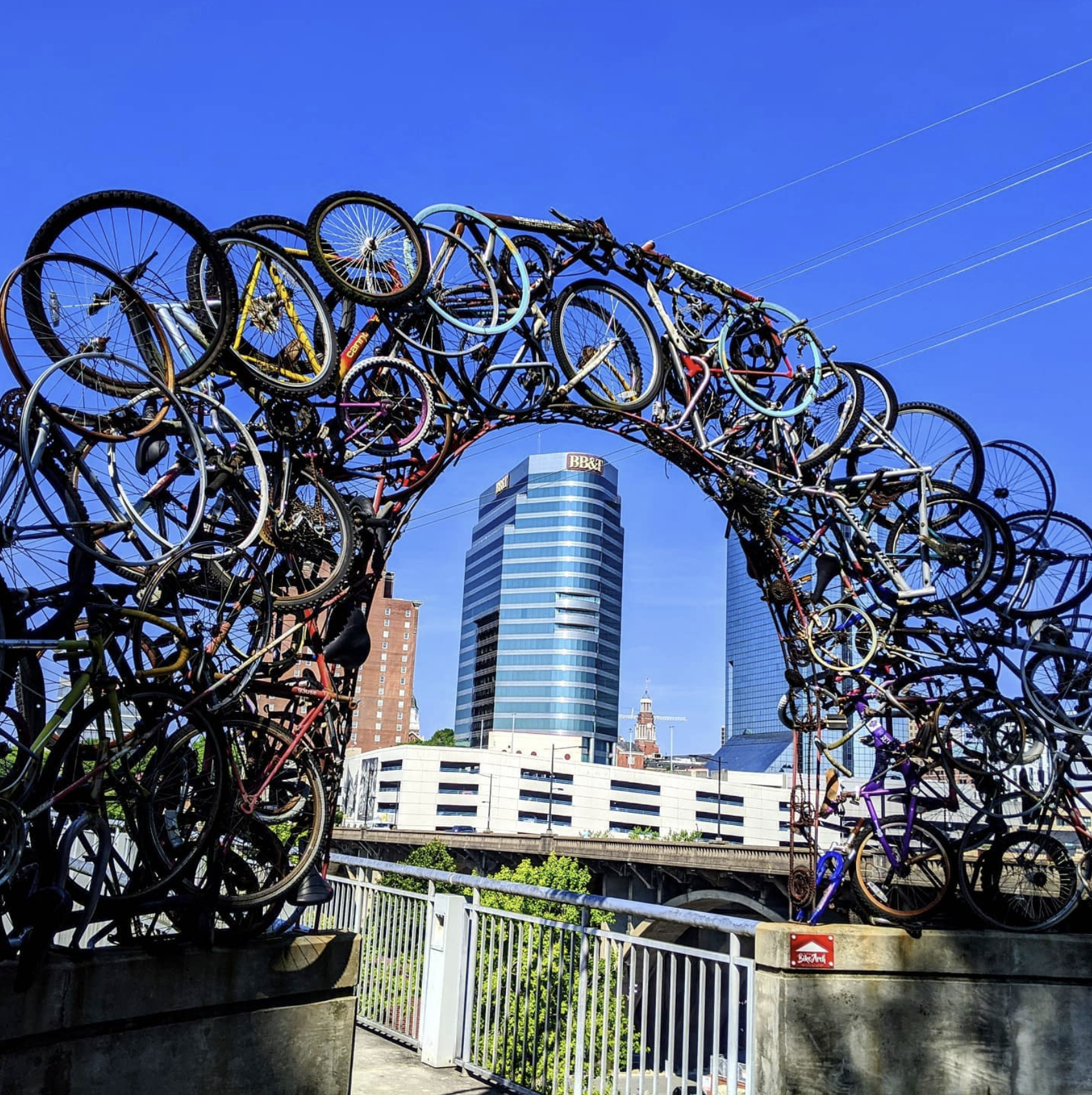 A bridge sculpture made of stacked and intertwined bicycles against a city skyline with modern high-rise buildings and a clear blue sky.