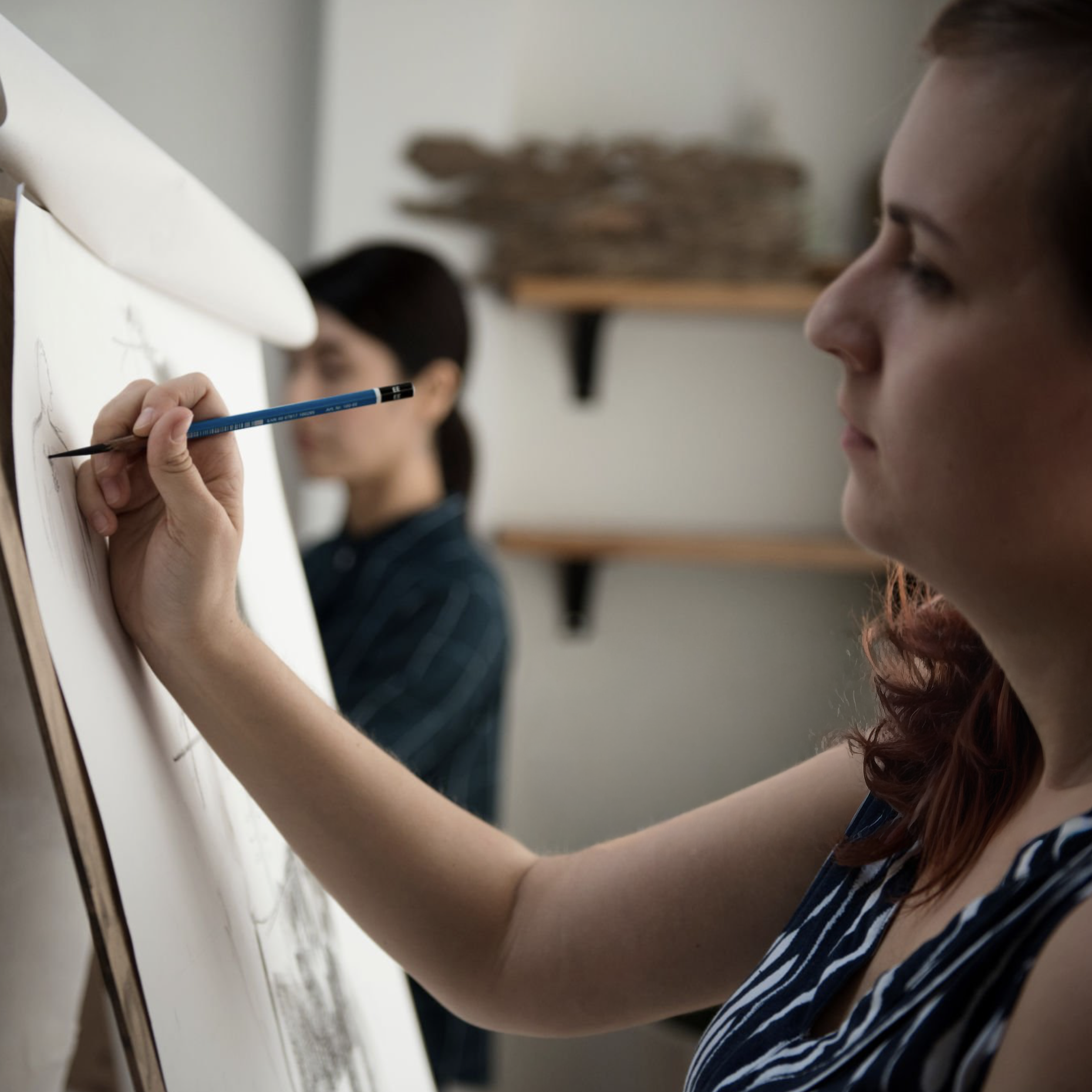 A woman drawing on a large sheet of paper with a pencil, focused on her artwork, in an art studio with shelves in the background.
