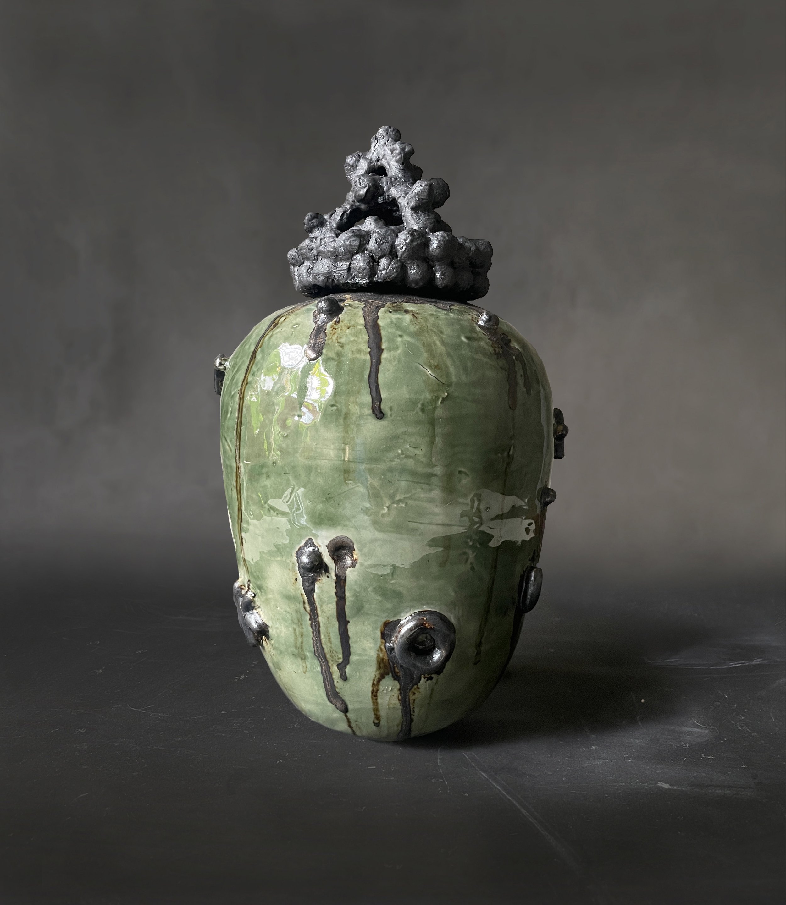 A green ceramic vase with black glaze drips and black clay-like protrusions, topped with a black, irregularly shaped sculpture.