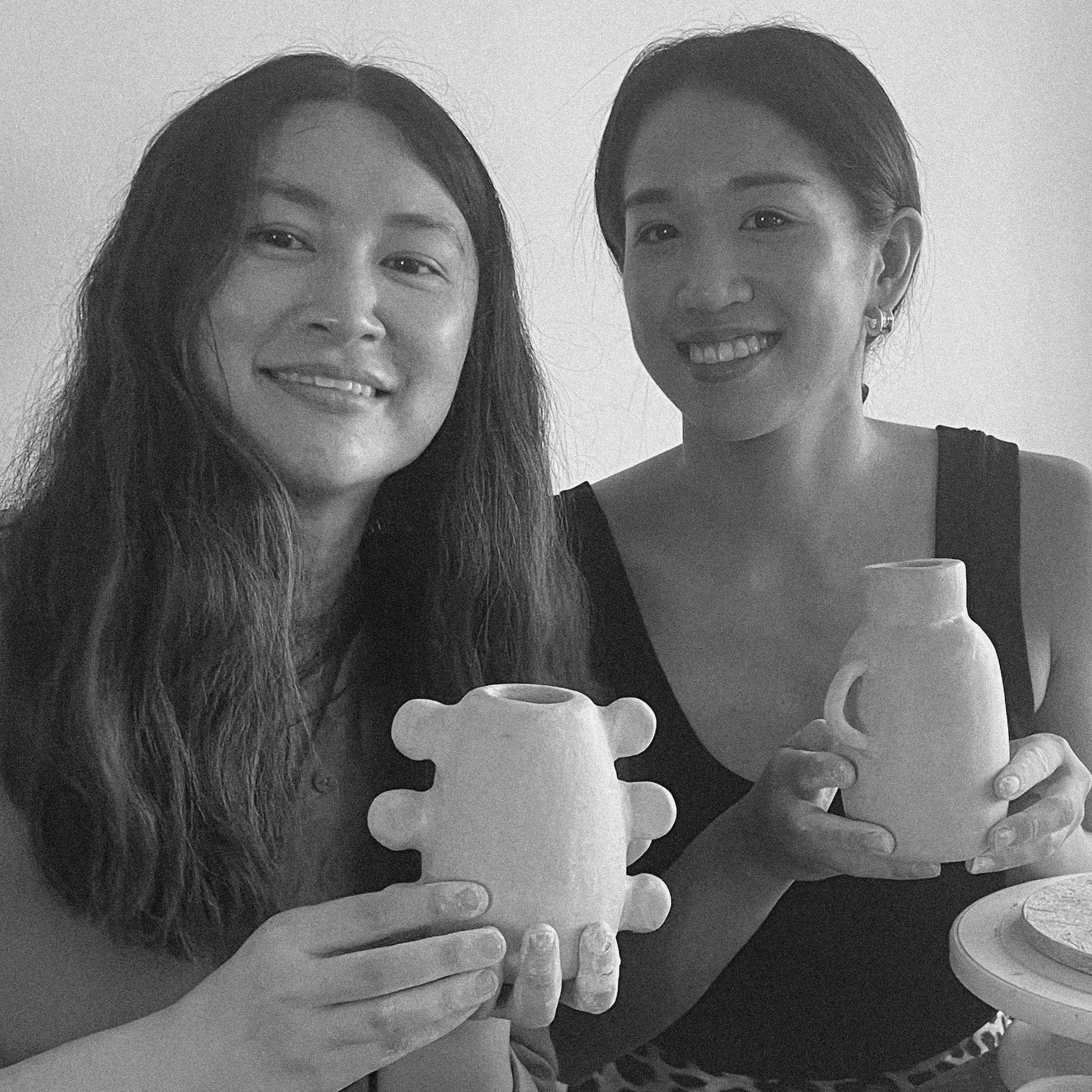 Two women smiling and holding ceramic vessels, standing against a plain wall, in a black-and-white photo.