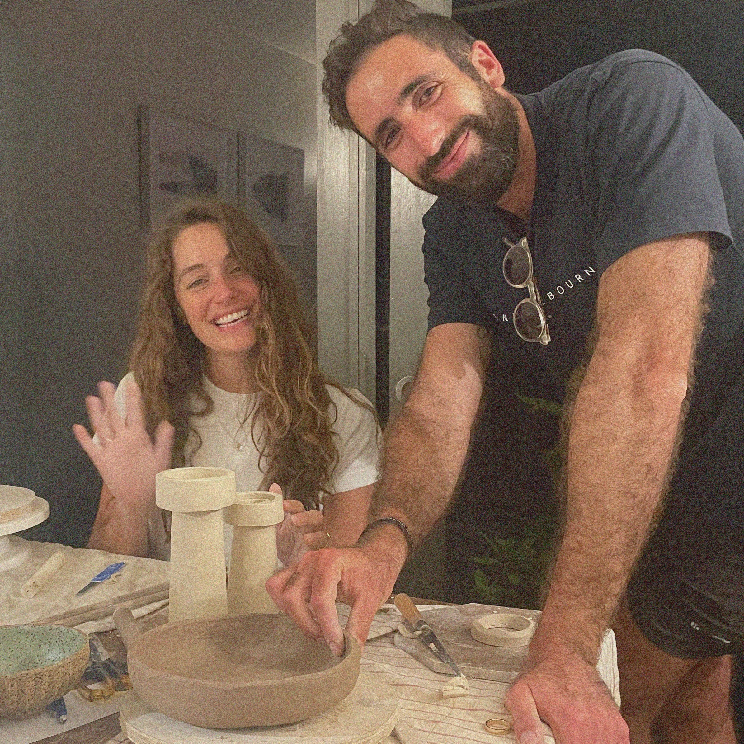 A man and a woman smiling at a pottery-making workshop. The man is shaping a clay bowl, and the woman is waving. The table is filled with pottery tools and clay.