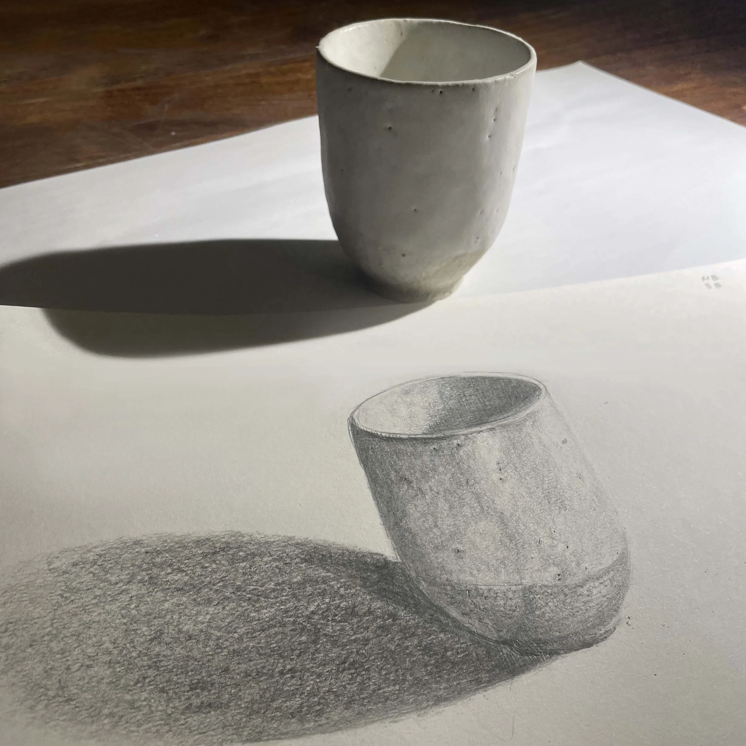 A ceramic cup on a paper surface, casting a shadow that appears as a detailed graphite pencil drawing of the cup.