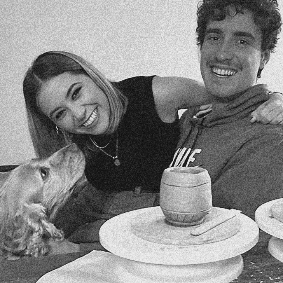 A smiling woman and man sitting at a table with a dog, plates, and a ceramic pot, in a cozy indoor setting.