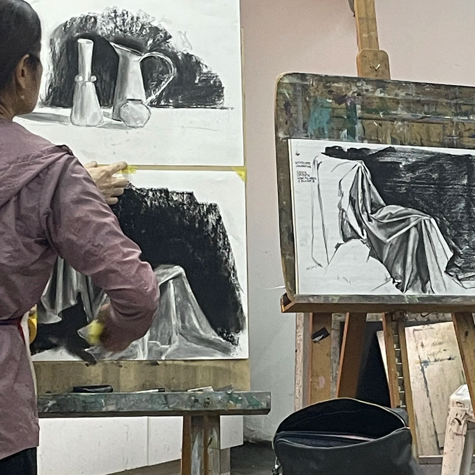 An artist creating a monochrome still life painting of vases on a table, with two canvases on easels showing different stages of the same scene.