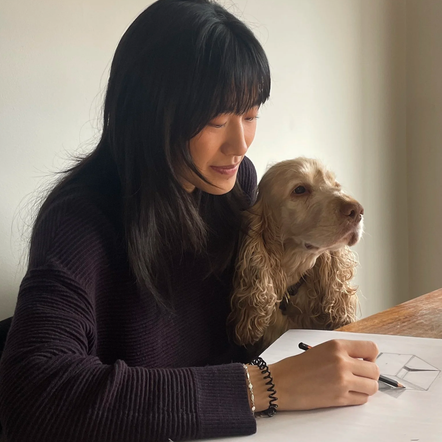 A woman with long dark hair sitting at a table, holding a pen, with a golden cocker spaniel dog sitting beside her. The woman is dressed in a dark long-sleeve shirt and appears to be writing or drawing on a sheet of paper.