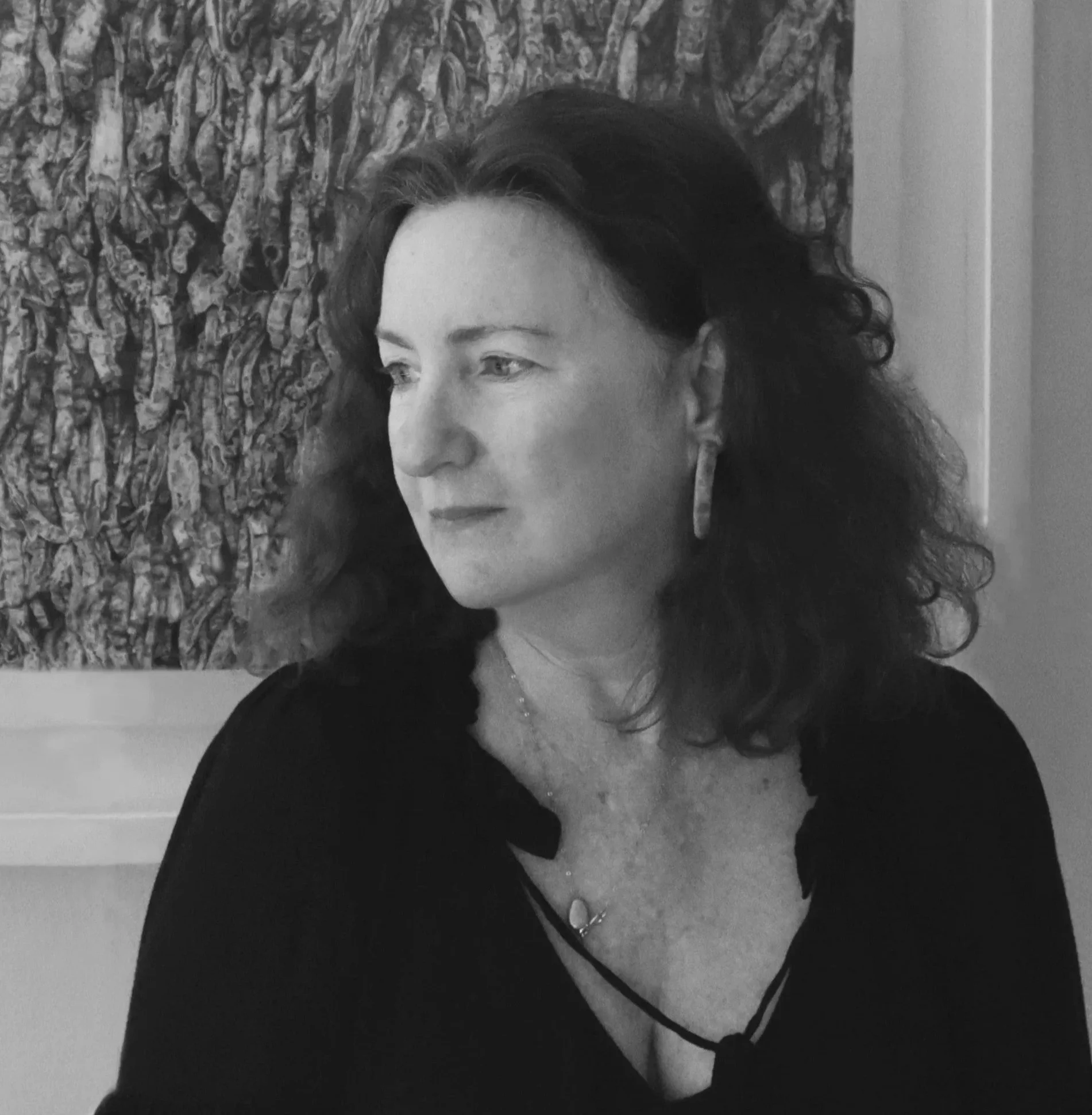 Black and white portrait of a woman with curly hair, wearing earrings and a necklace, looking to her left, standing in front of a textured abstract art piece.