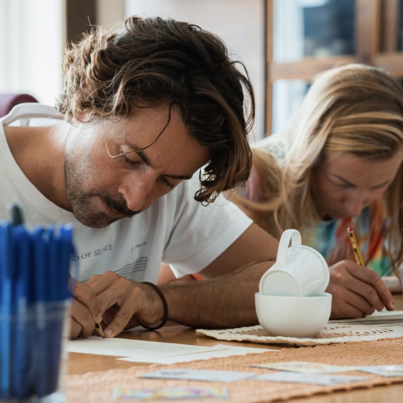 Two adults sketching or drawing at a table with art supplies, including a white teapot and cups