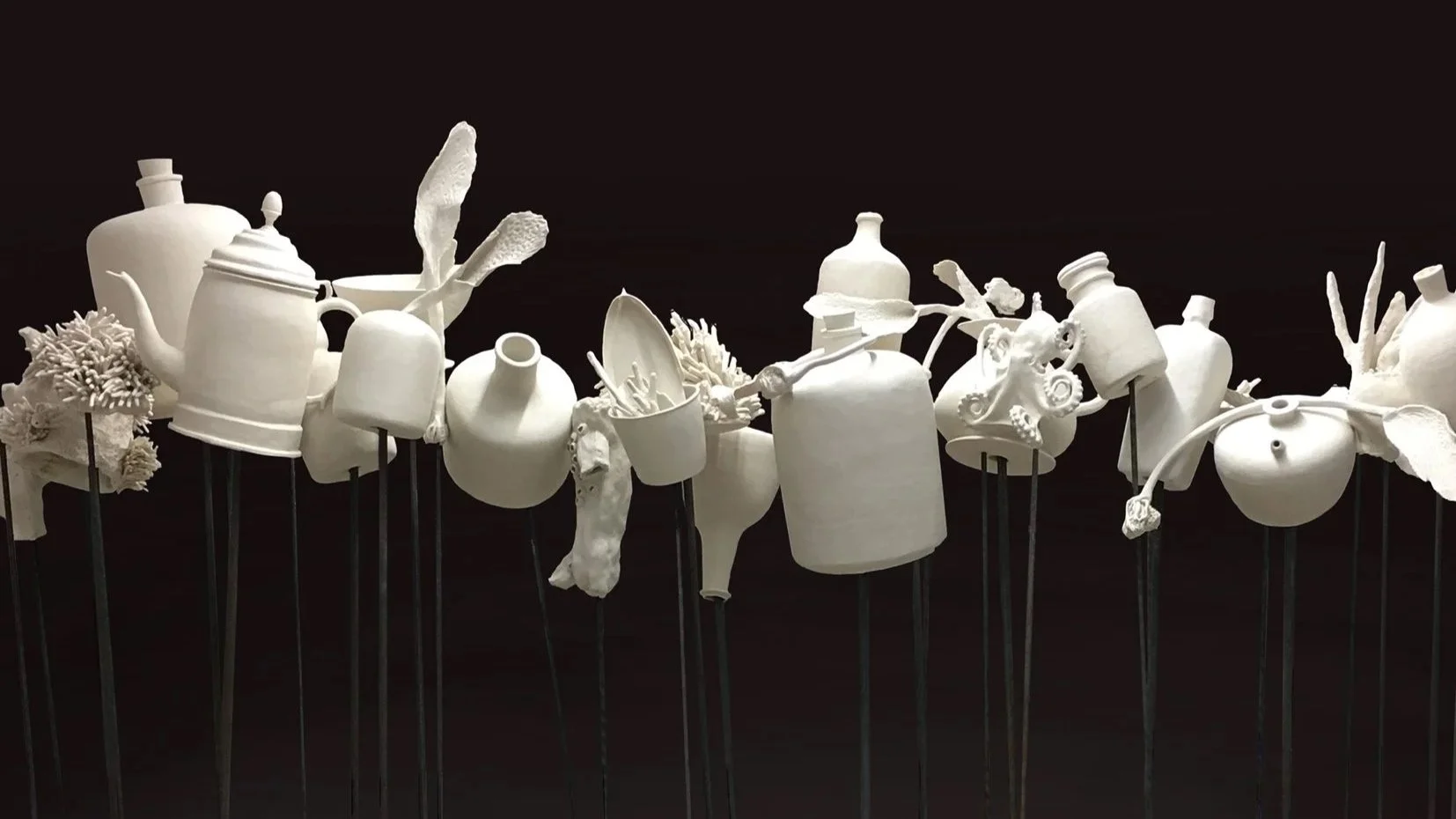 White ceramic or porcelain sculptures resembling various objects and forms mounted on black support rods against a black background.