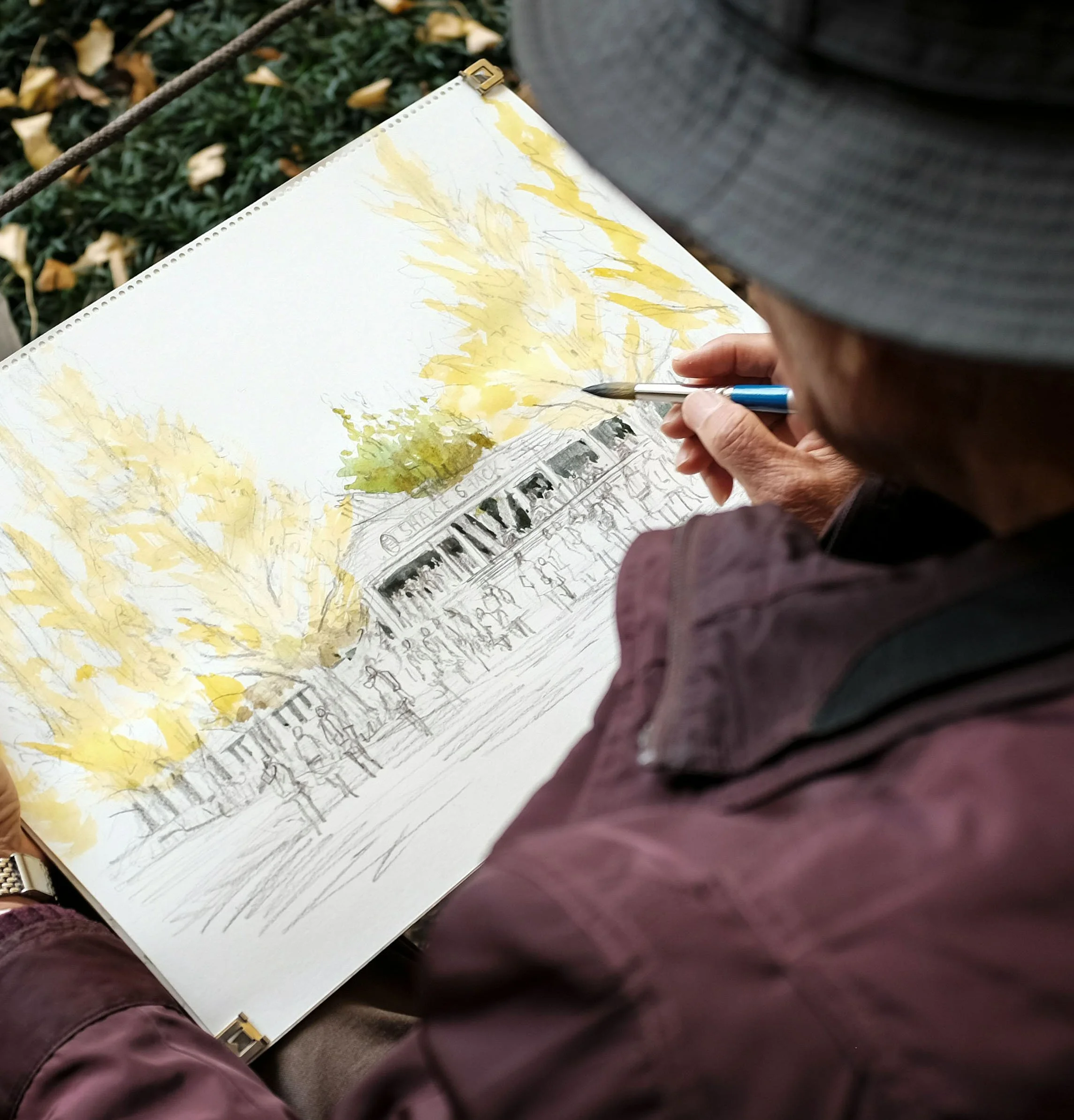 Person drawing a sketch of a crowded outdoor scene with trees and a building, using a pen on paper.