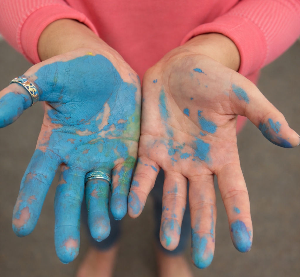 Hands covered in blue and pink paint, wearing rings, with a person in a pink sweater in the background.