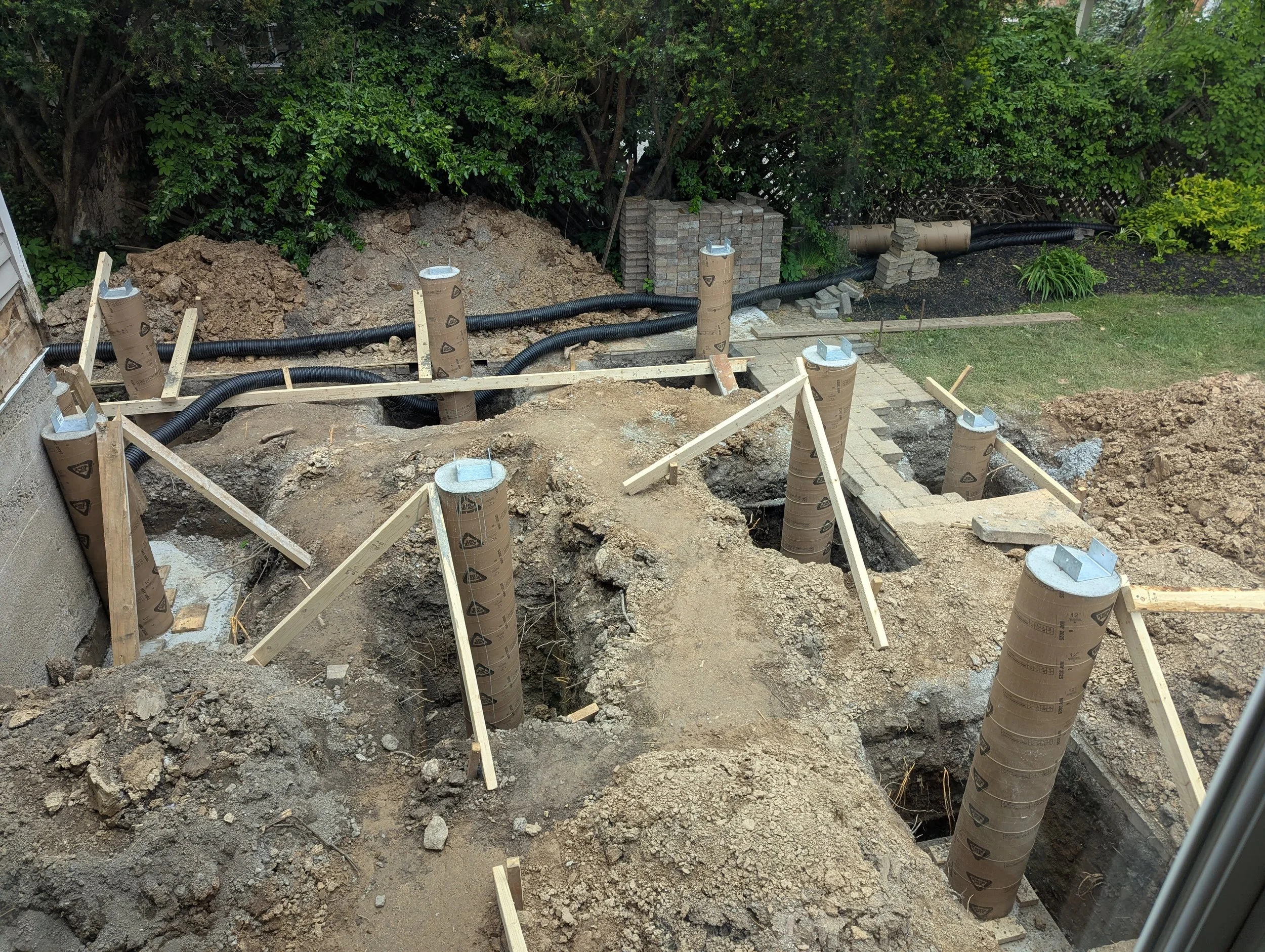 Construction site with concrete piers and black pipes, surrounded by dirt and wooden supports, in a backyard with greenery and brickwork.