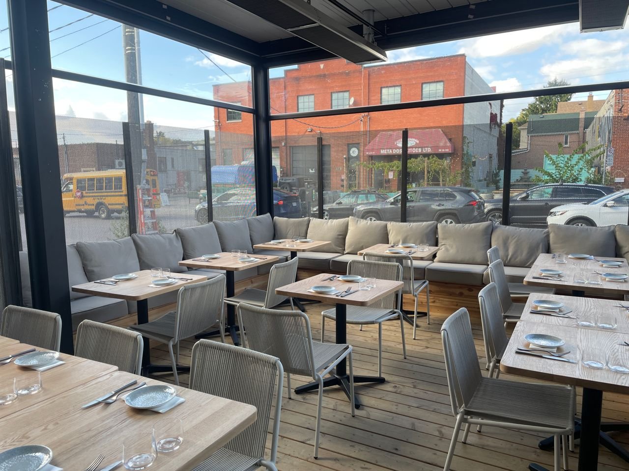 Empty restaurant dining area with wooden tables, metal chairs, and a long couch along the windows, with a view of the street outside.
