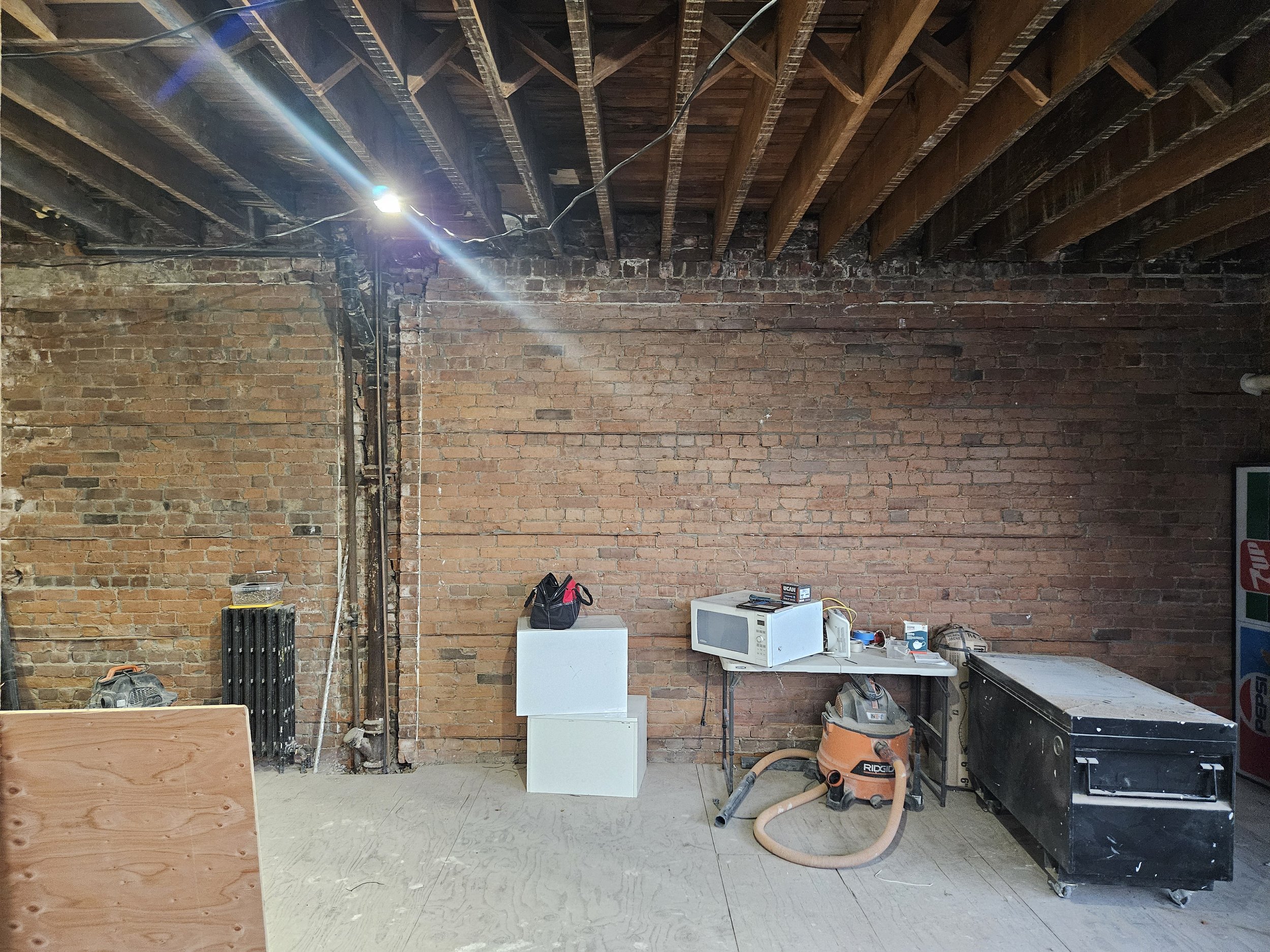 Interior of a basement or garage with exposed brick walls and unfinished ceiling beams. Contains a microwave, vacuum, chest freezer, and miscellaneous items on a table, with plywood panels and a Pepsi vending machine partially visible.