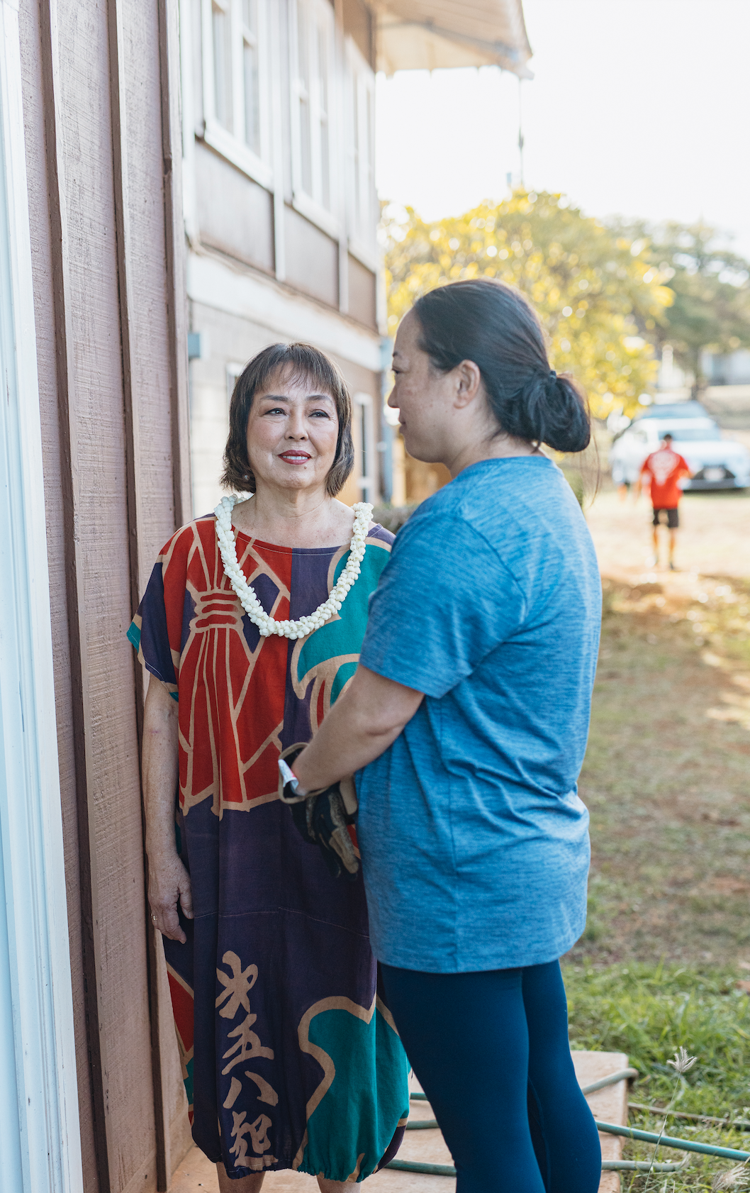 Two women engage in a conversation outdoors near a wooden building, one wearing a colorful dress and a white lei, the other in a blue shirt, with trees and cars in the background.