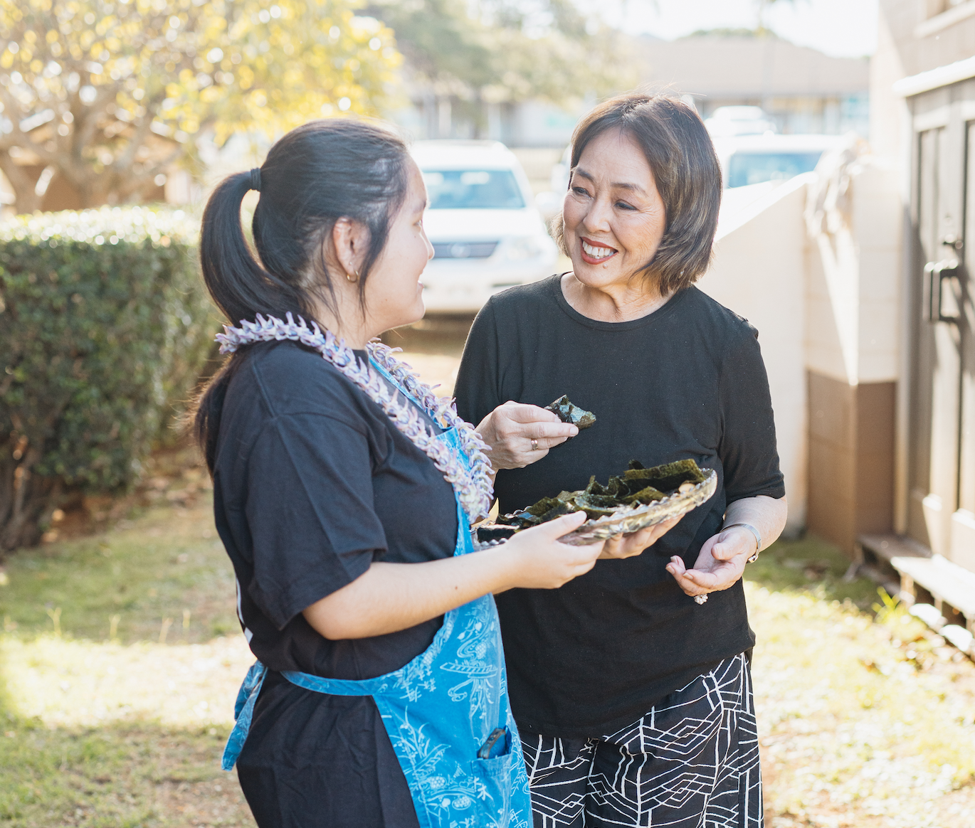 Two women, one wearing a black shirt and patterned pants, and another in a black shirt and blue apron, are talking outdoors. One woman is holding a plate of seaweed snacks and offering a piece to the other woman, who is smiling.