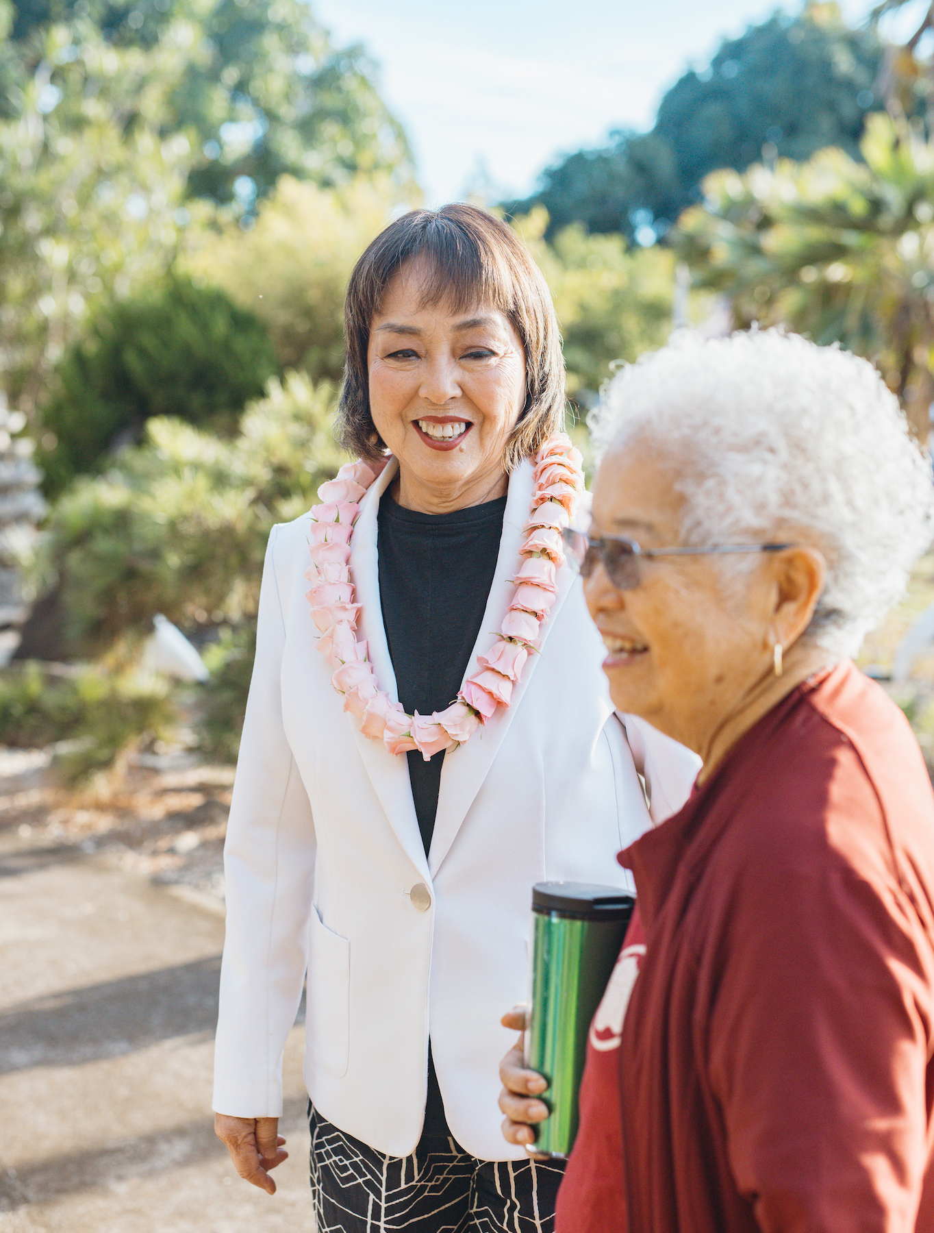 Two women smiling outdoors; one wearing a white blazer with a pink lei, the other in a maroon jacket holding a green tumbler.