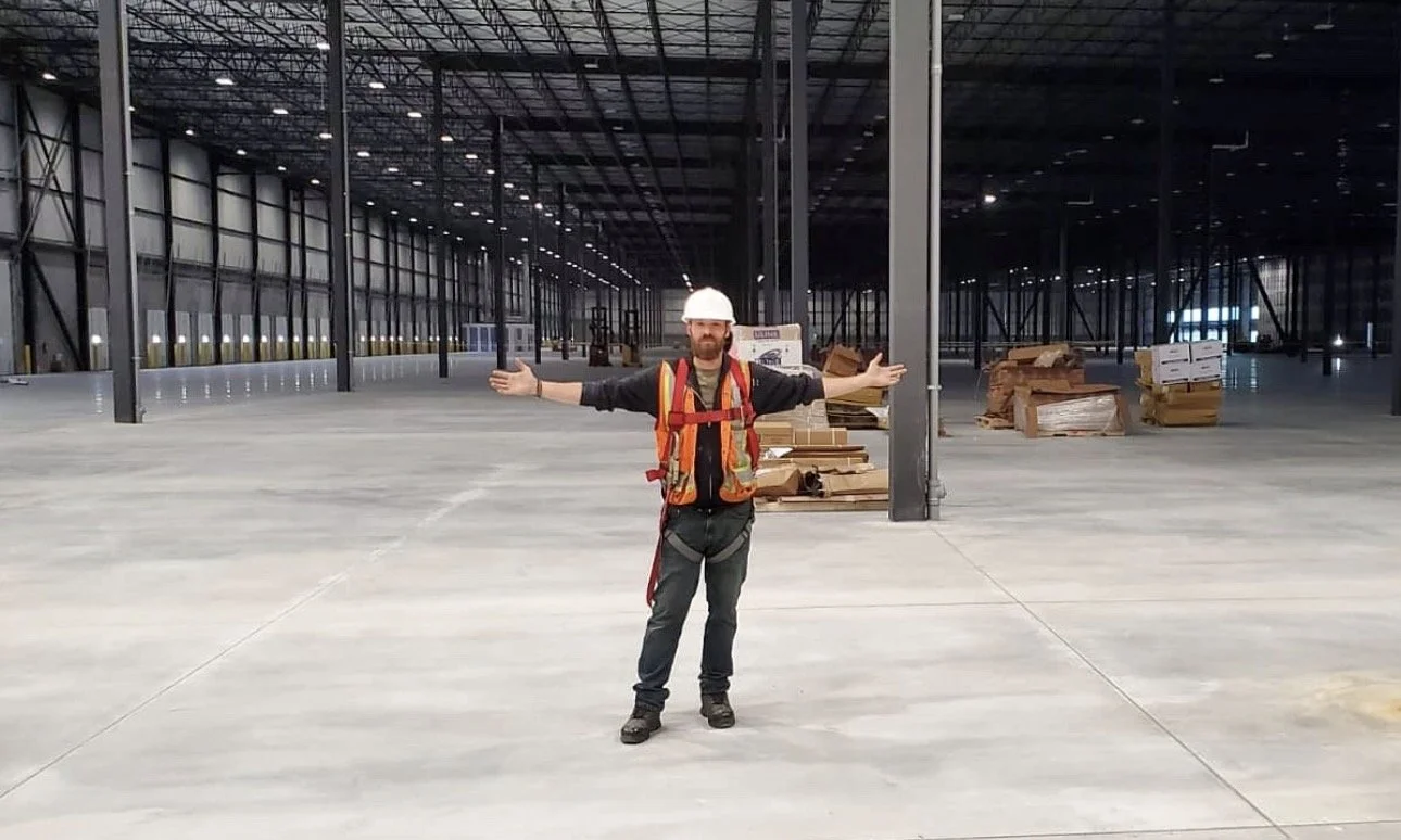 Construction worker standing with arms wide open inside a large, empty warehouse with high ceilings and exposed steel beams.