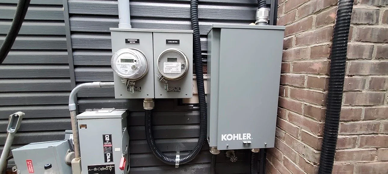 Electrical meters, a gray KOHLER electrical box, and various wiring mounted on a brick and metal exterior wall.