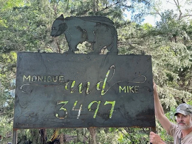 A man holding a large metal sign with a bear on top and an engraved name 'Monique and Mike 3497' in a wooded area.