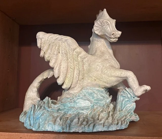 A sculpture of a winged horse, resembling Pegasus, made of light-colored material on a wooden shelf.