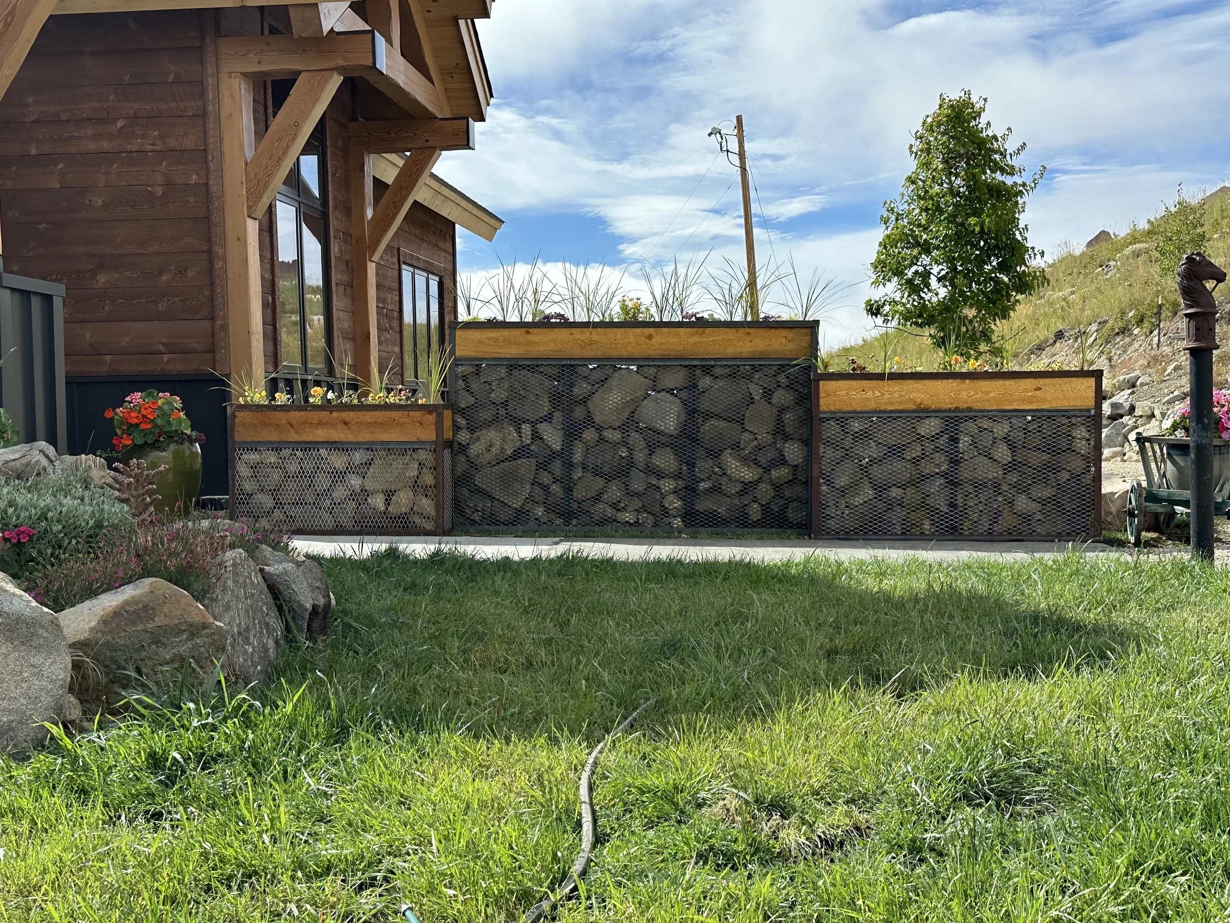 A landscaped yard with a wooden house, a rock-filled gabion wall planter, colorful flowers, green grass, a small tree, a hill with rocks, and a cloudy sky.