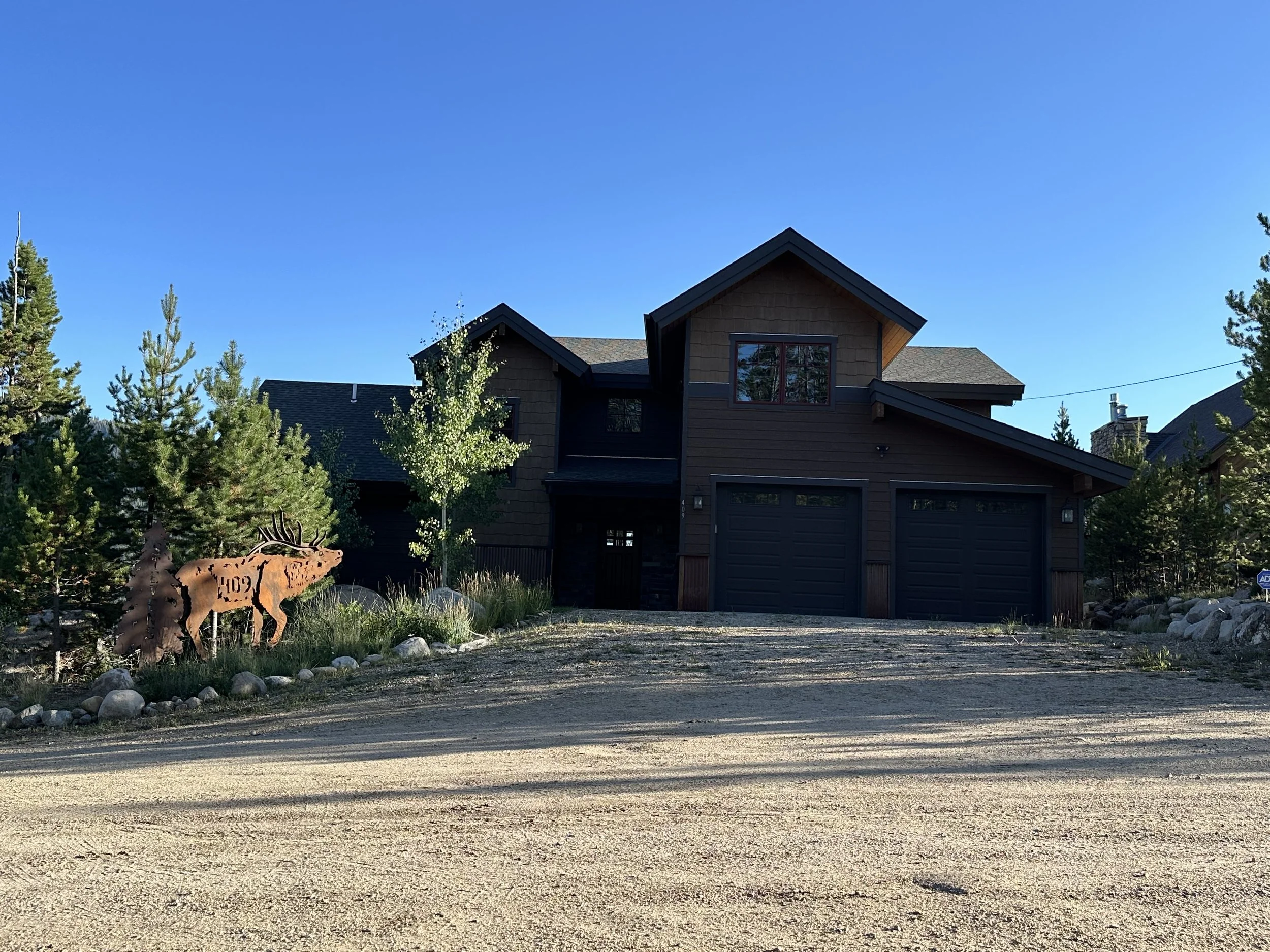 A modern two-story house with a three-car garage, dark exterior, and a dirt driveway. There are trees and landscaping around the house, and a wooden moose sculpture in the yard.
