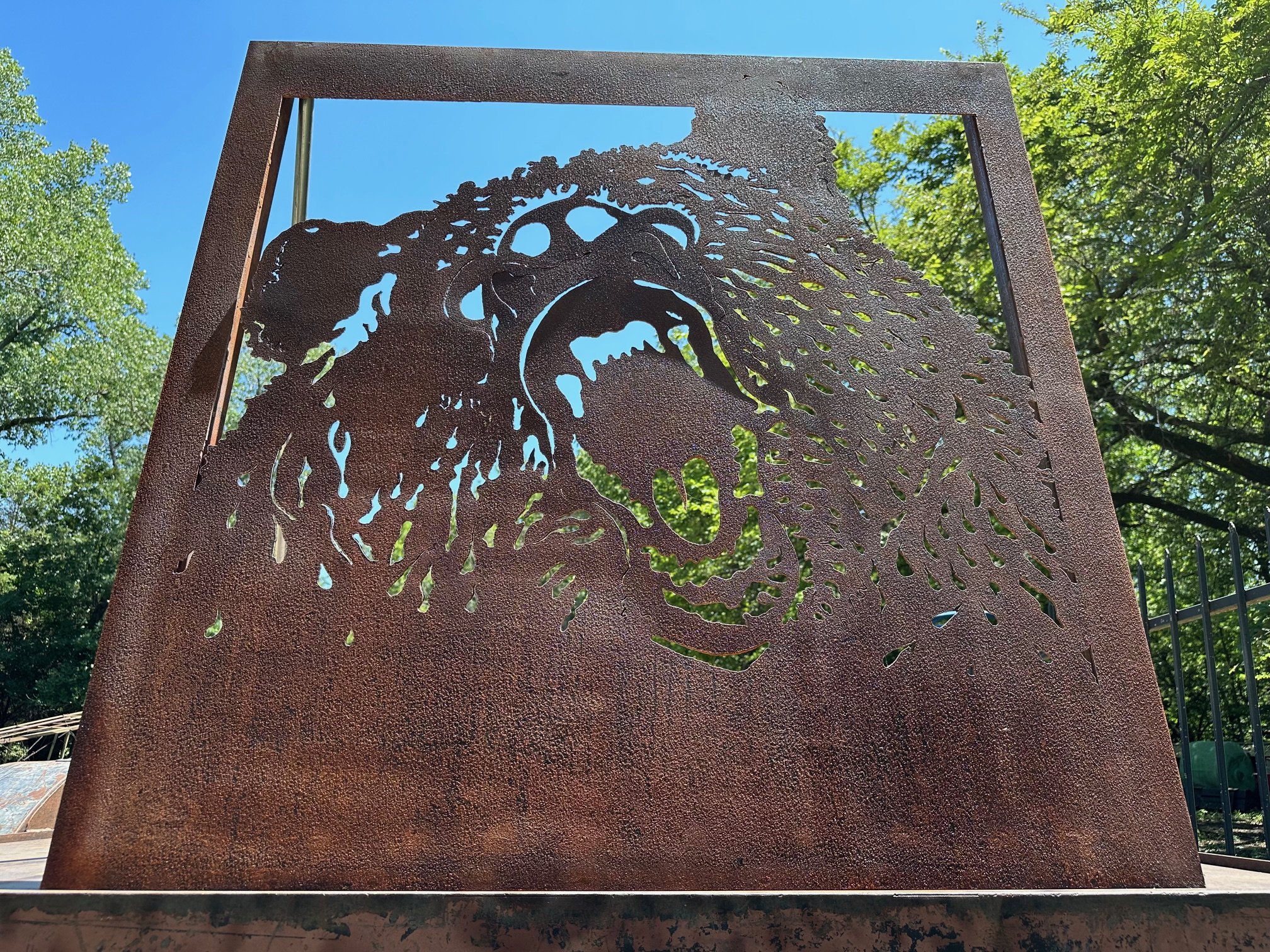 Metal sculpture of a roaring bear's head with detailed cutout design, set outdoors against green trees and a blue sky.