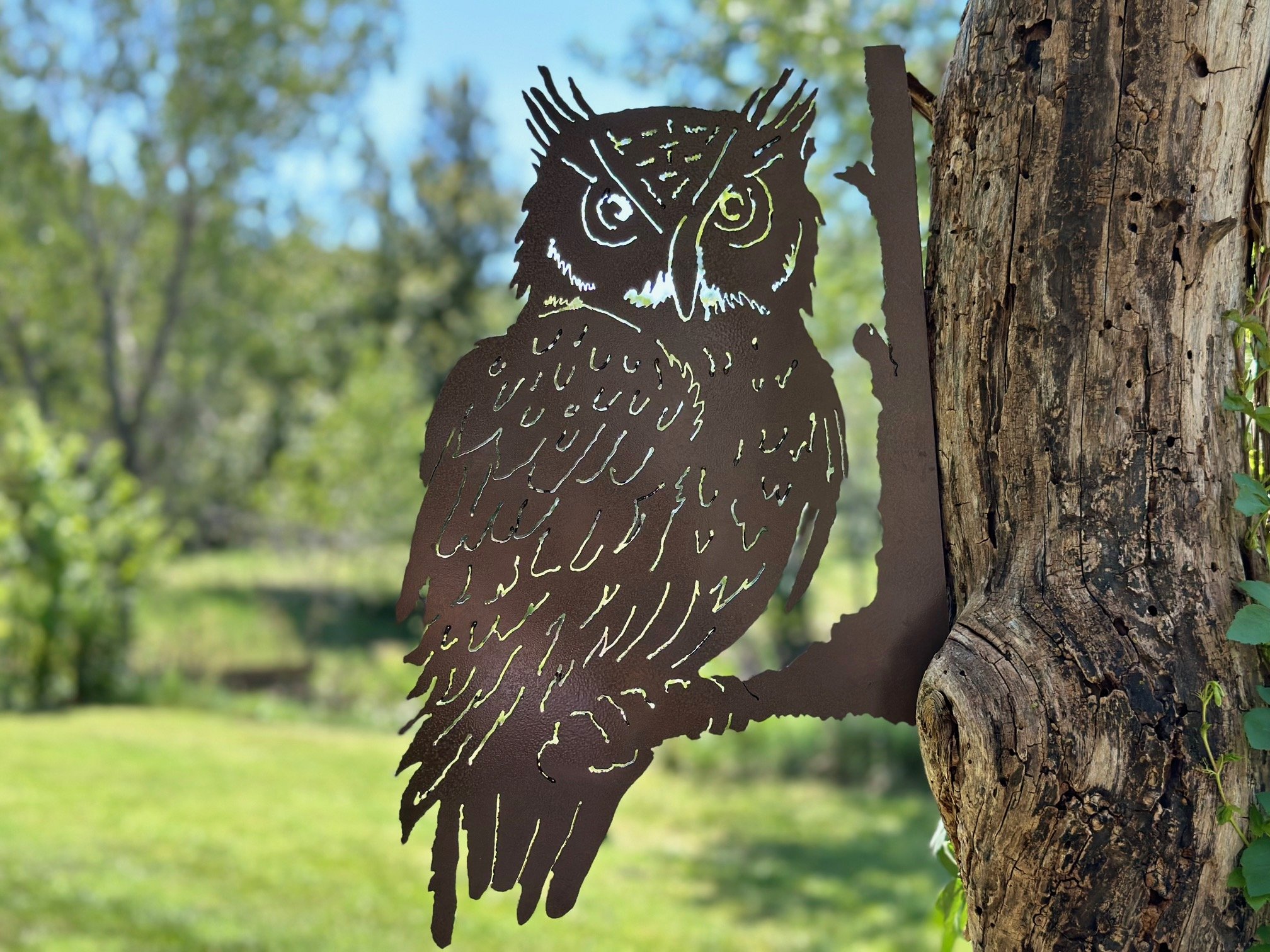 Metal cutout of an owl mounted on a tree trunk in a forested area with green trees and grass in the background.