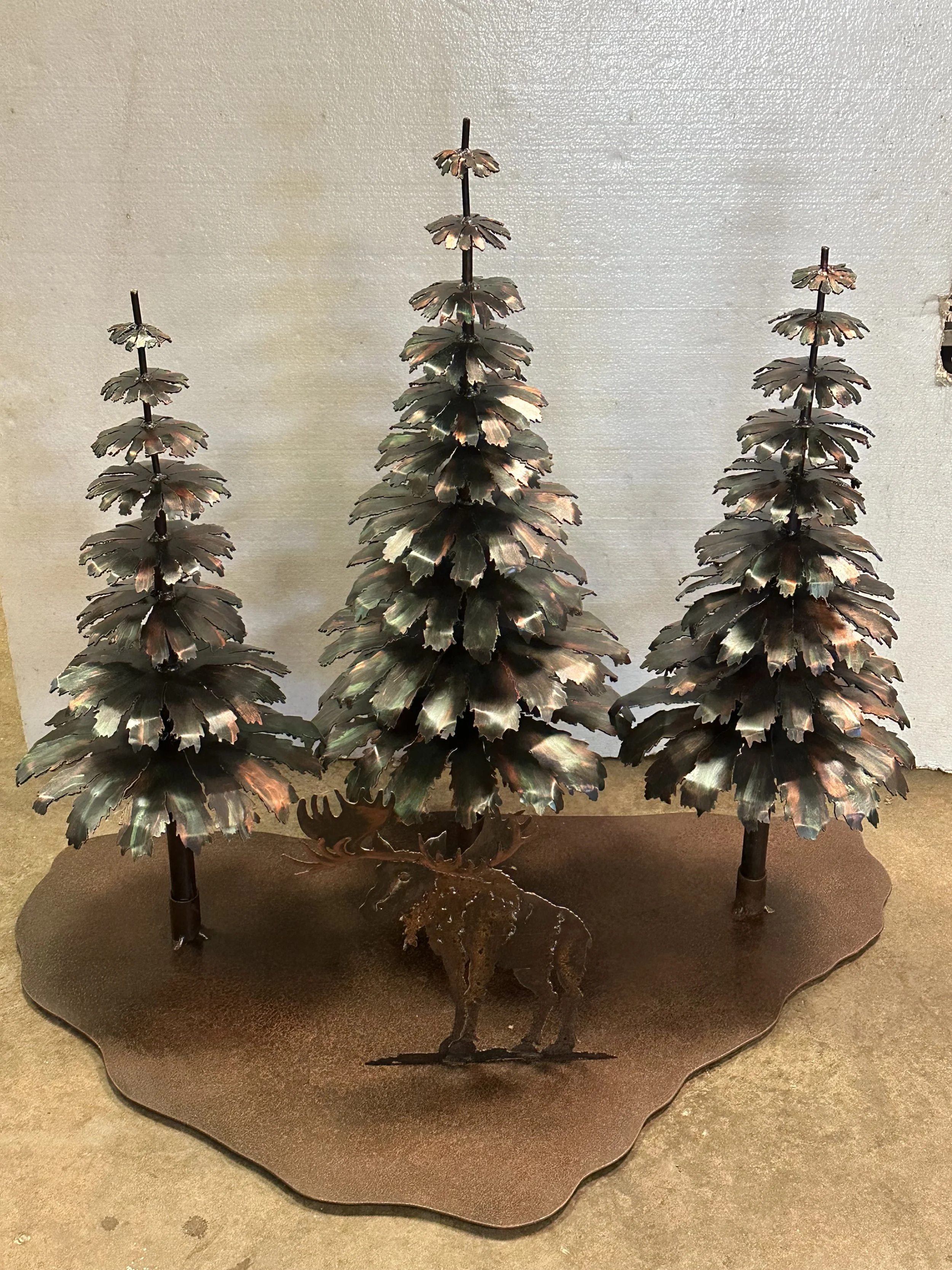 Metal sculpture of three stylized evergreen trees with large, layered leaves, mounted on a base with a textured surface.
