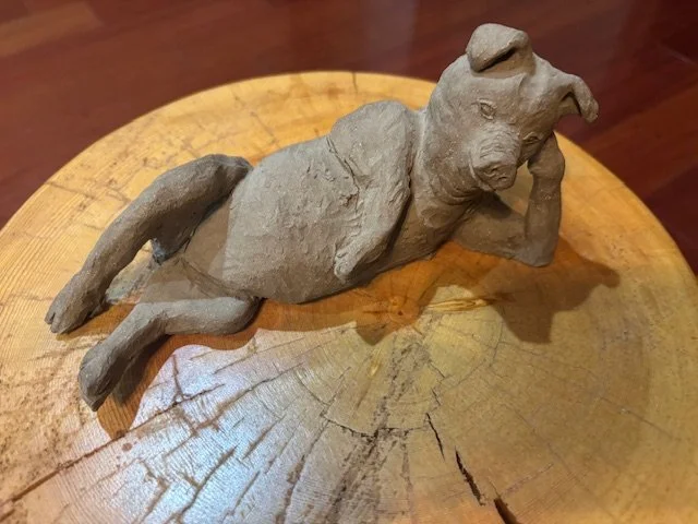 A small sculpture of a dog lying on its side with one paw supporting its head, made of clay or similar material, placed on a wooden surface.