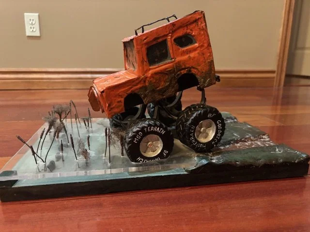 A model of an orange off-road vehicle with large black wheels positioned on a small terrain with miniature trees, placed on a black base on a wooden floor.