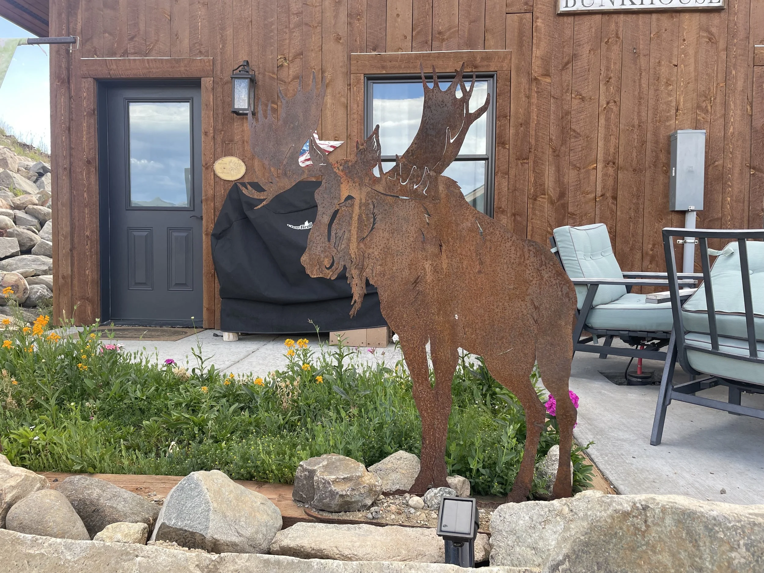 Rusty metal sculpture of a moose outdoors in front of a wooden building with patio furniture and a flower garden.