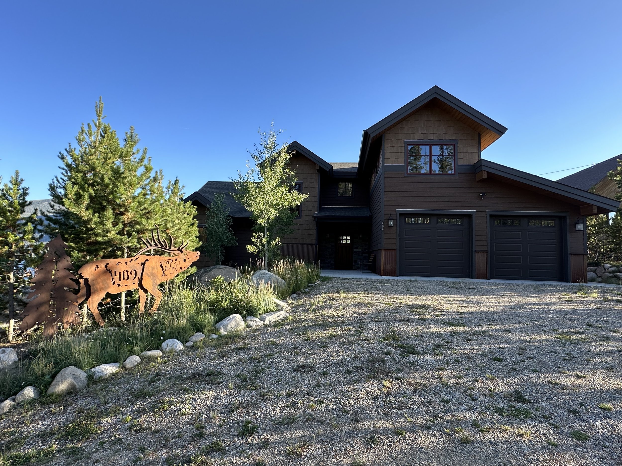 Front view of a two-story house with a brown exterior, black garage doors, and a grey roof under a clear blue sky. There are trees and large rocks in the yard, with a metal moose yard decoration in the foreground.