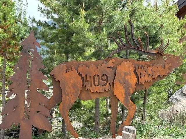 Rusty metal art sculpture of a moose with antlers, set against trees and a wooden cabin.