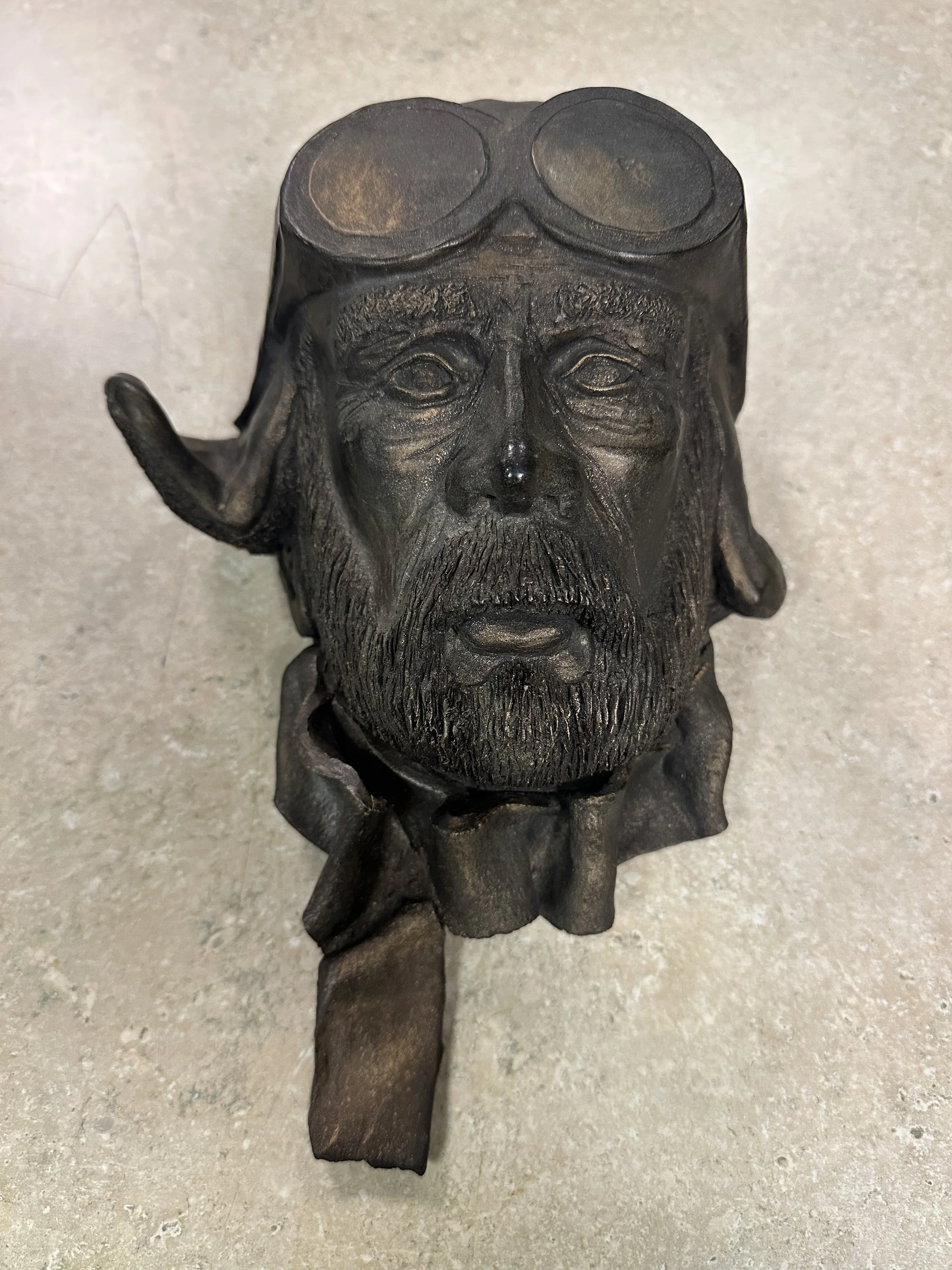 A dark metallic statue of a bearded man with long hair and a serious expression, with sunglasses on his head, and a small figure at the base, set on a light-colored surface.