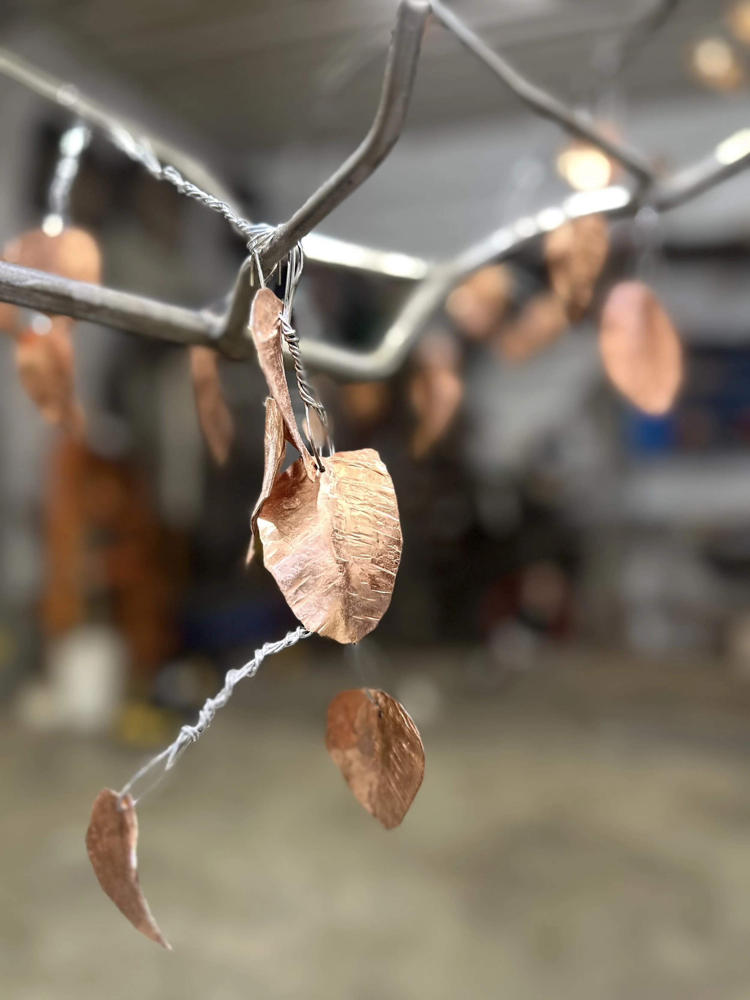 Close-up of copper leaves hanging from a wire on a metallic branch, blurred workshop background.
