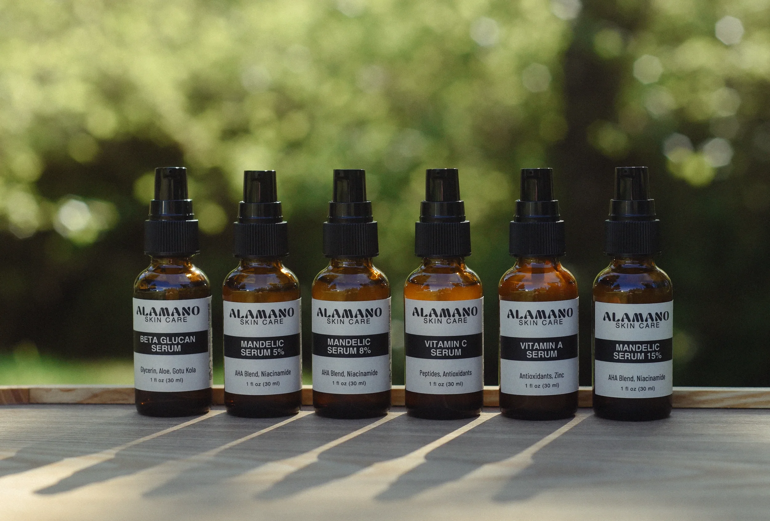 Six small brown glass bottles with black spray caps, labeled with various skin care serums from Alamano. The labels indicate ingredients such as Beta Glucan, Mandelic Acid, Vitamin C, Vitamin A, and other components. The bottles are arranged in a line on a wooden surface with a blurred green forest background, and shadows cast on the surface.