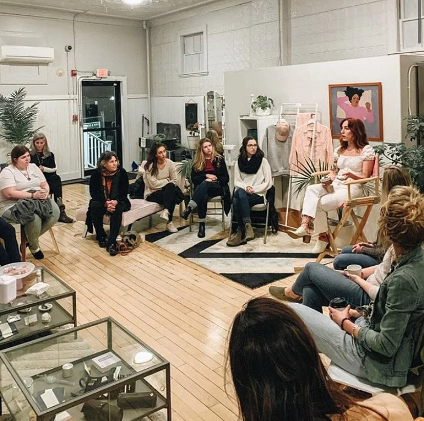 A group of women seated in a semi-circle in a cozy, decorated room, listening to a woman in white speaking.