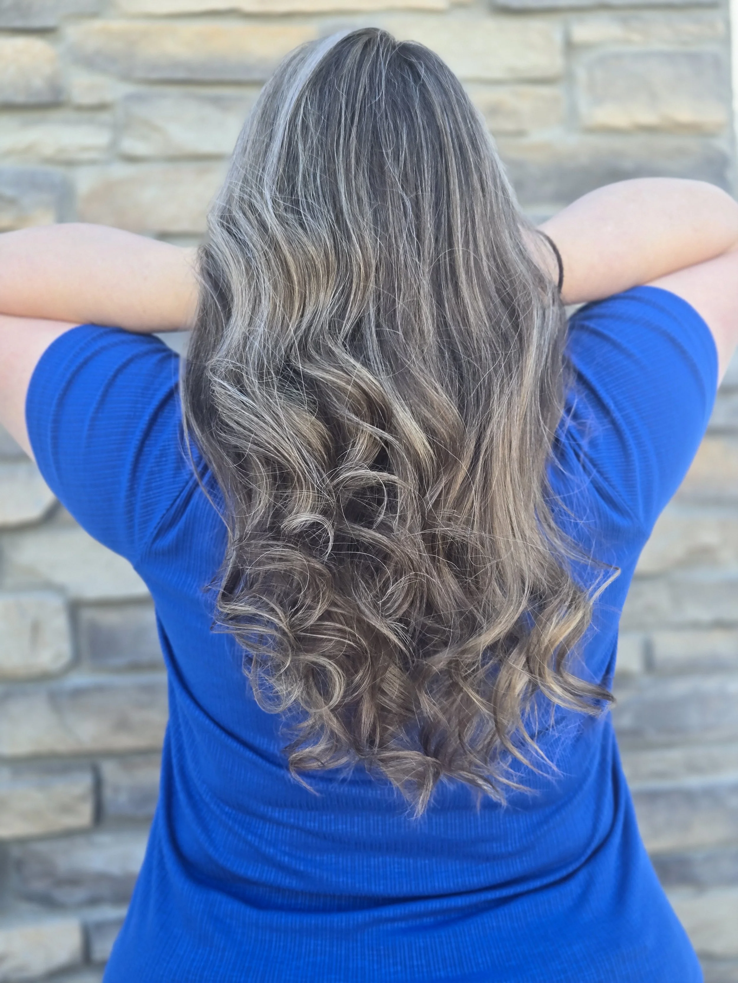 Back view of a woman with long, wavy, light brown hair wearing a blue shirt, standing against a stone wall.