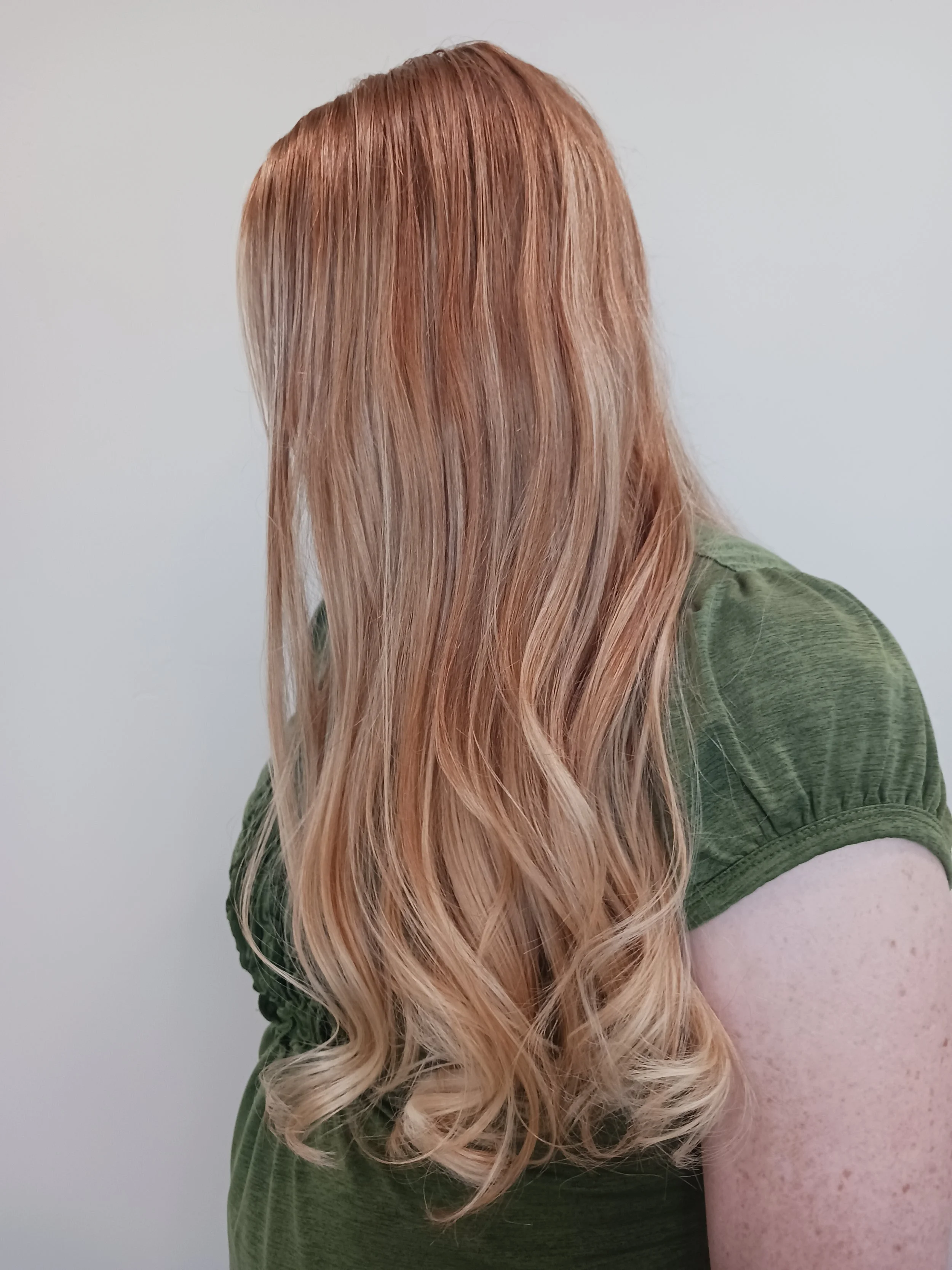 A person with long, wavy, blonde hair wearing a green shirt, facing away against a plain light background.