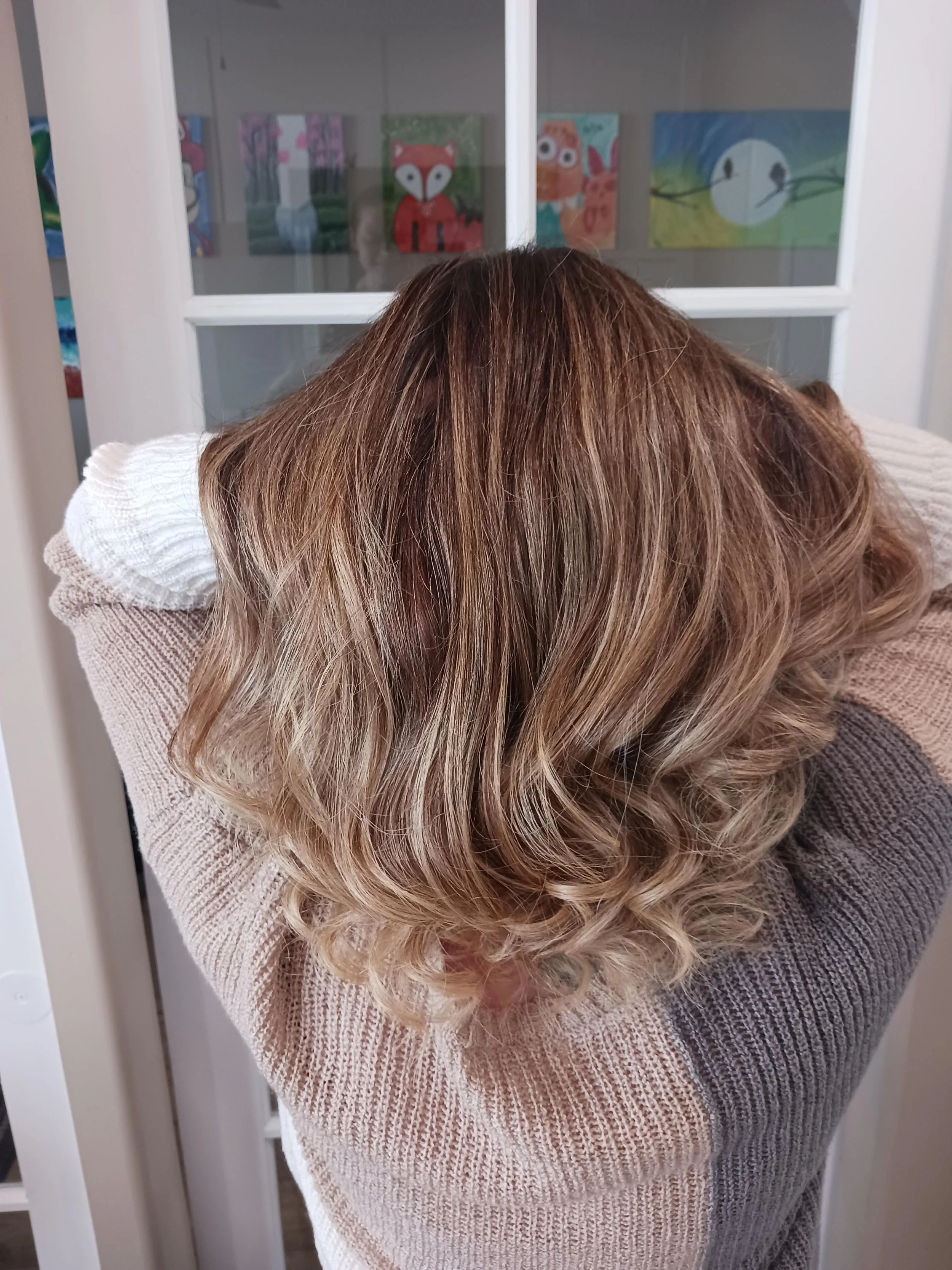 Back view of a woman with shoulder-length wavy blonde hair, resting her head on her crossed arms, standing in front of a glass door with children's artwork visible through the glass.
