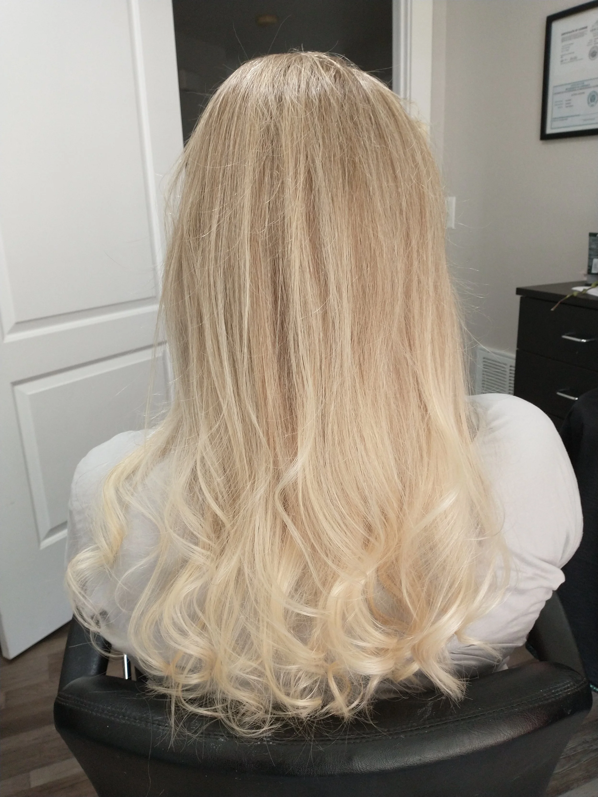 Back of a woman with long, wavy blonde hair, sitting in a chair in what appears to be a professional setting with framed certificates on the wall.