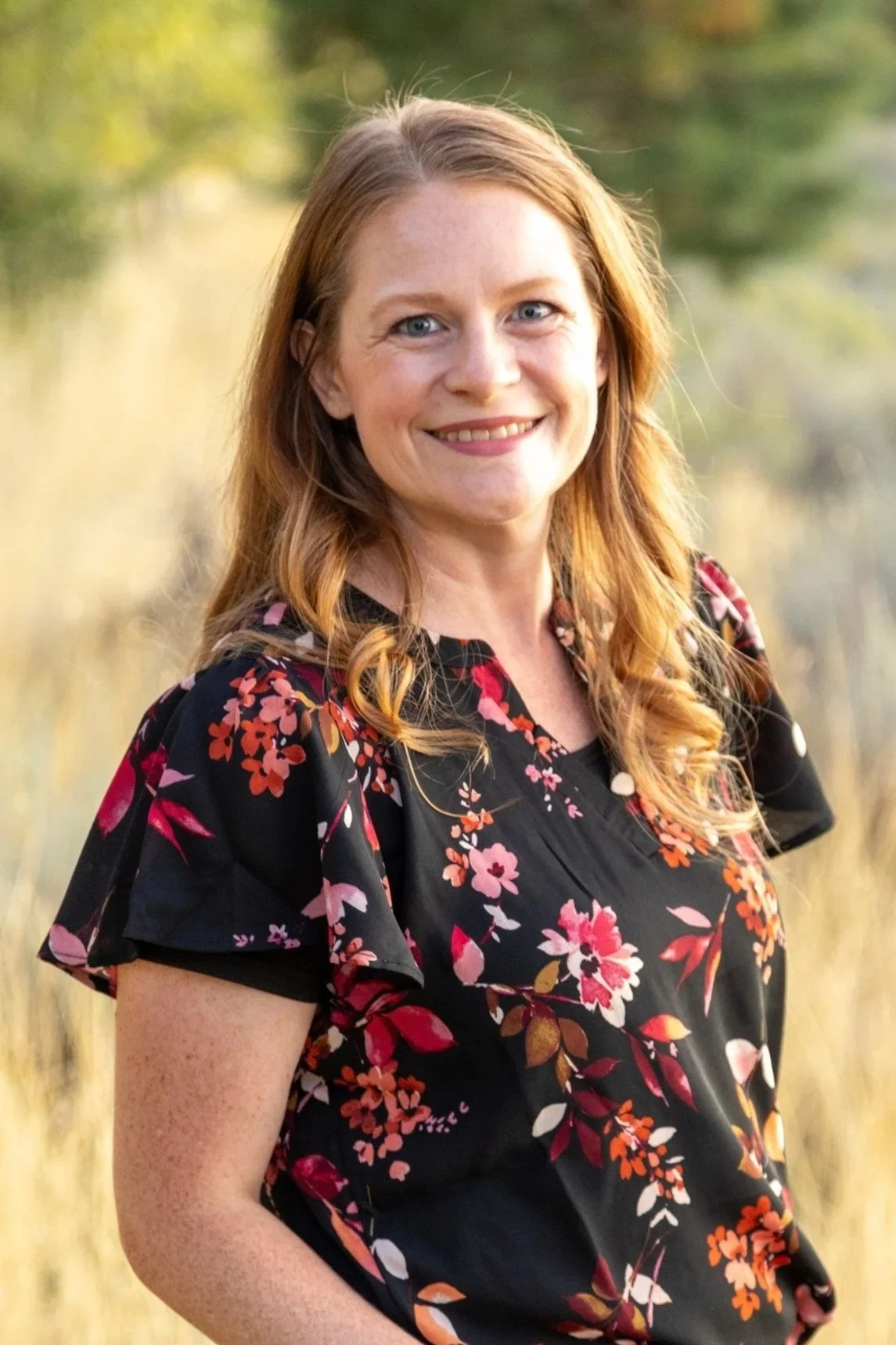 A woman with red hair and blue eyes smiling outdoors, wearing a black dress with a red, pink, and white floral pattern, with a blurred natural background.