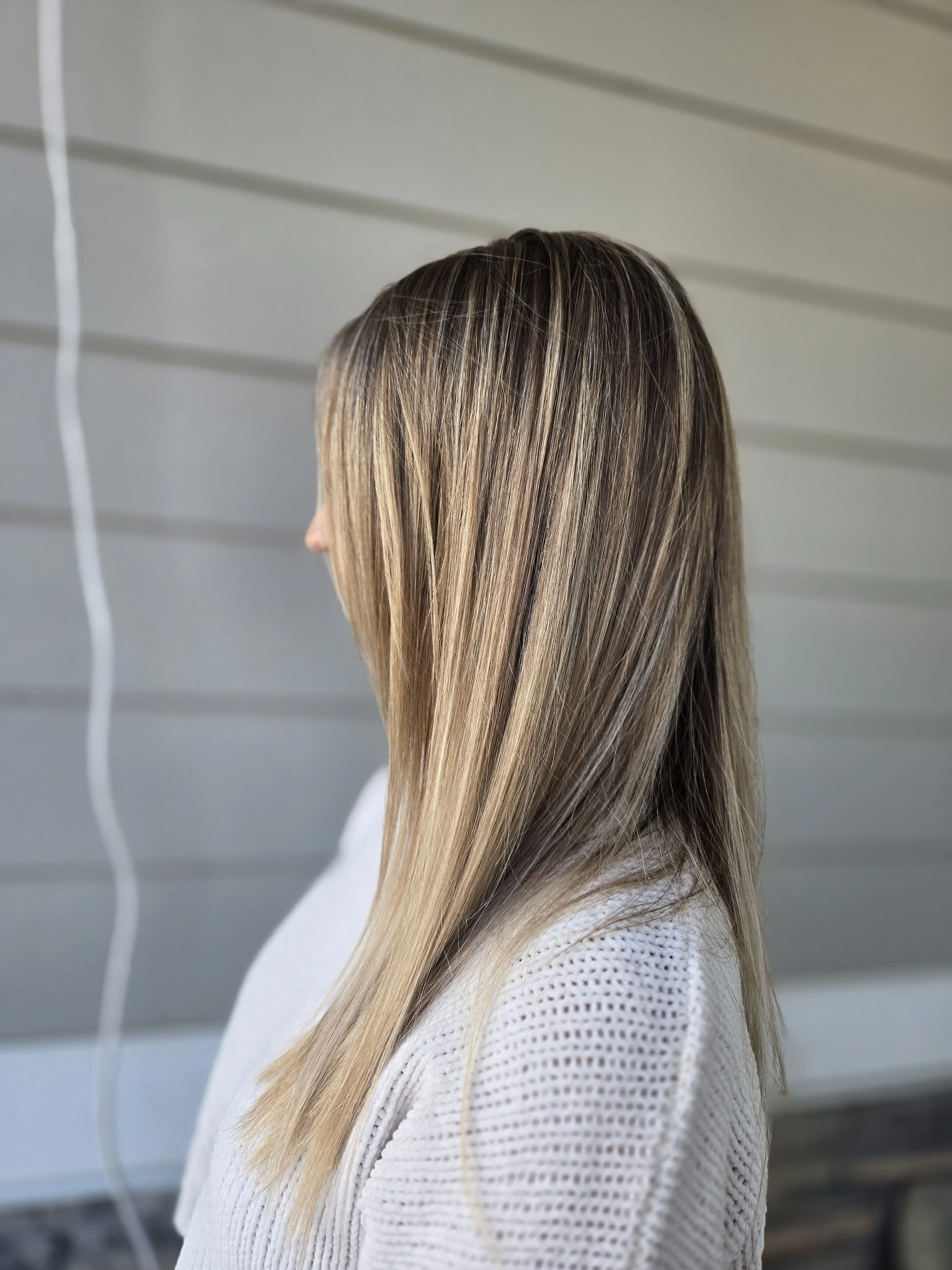 A woman with straight, shoulder-length hair with blonde highlights, wearing a white knit sweater, standing sideways with a gray siding background.