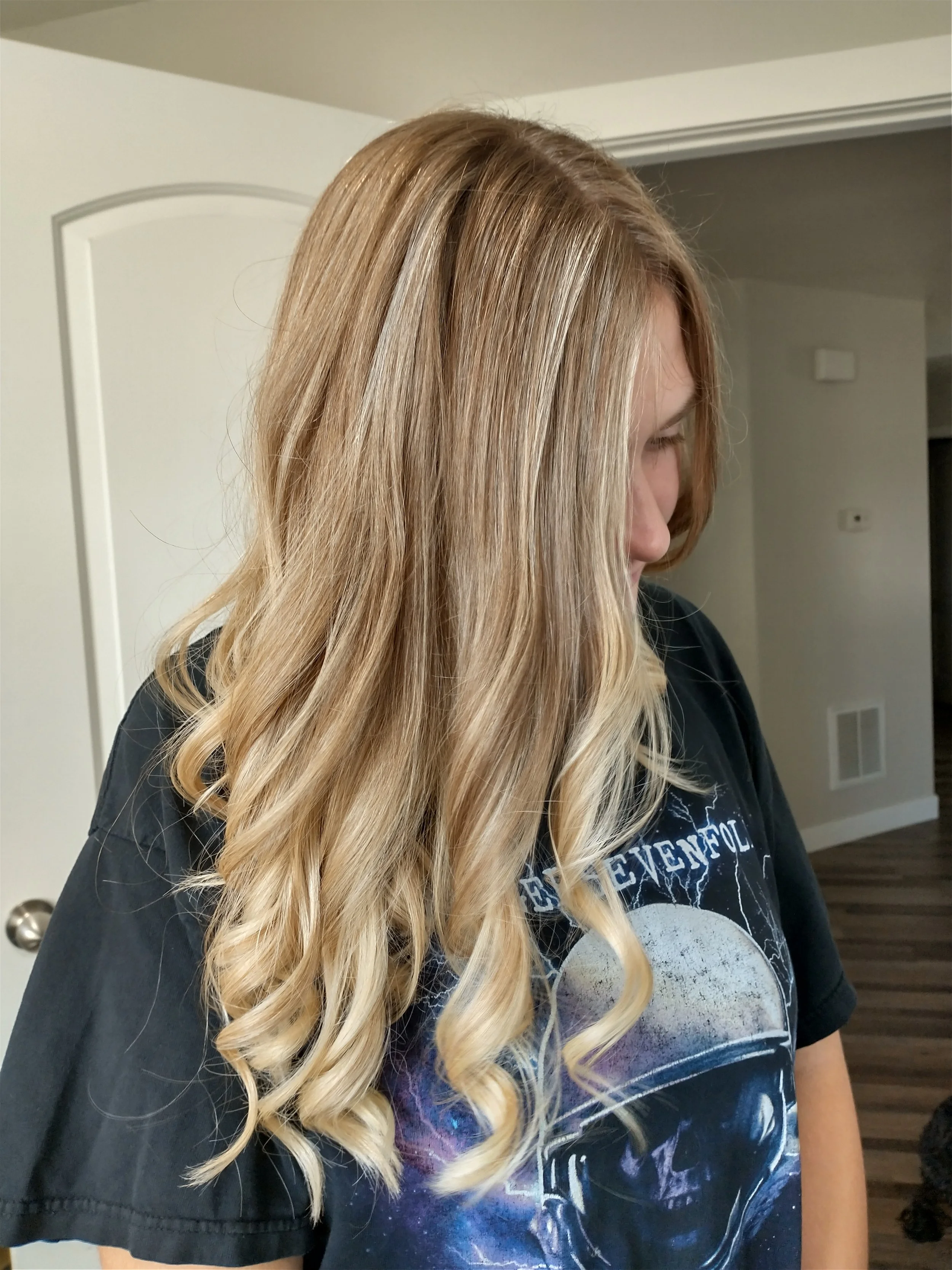 A woman with long, blonde, wavy hair, looking down, wearing a black T-shirt with a graphic design, standing in a room with beige walls and a wooden floor.
