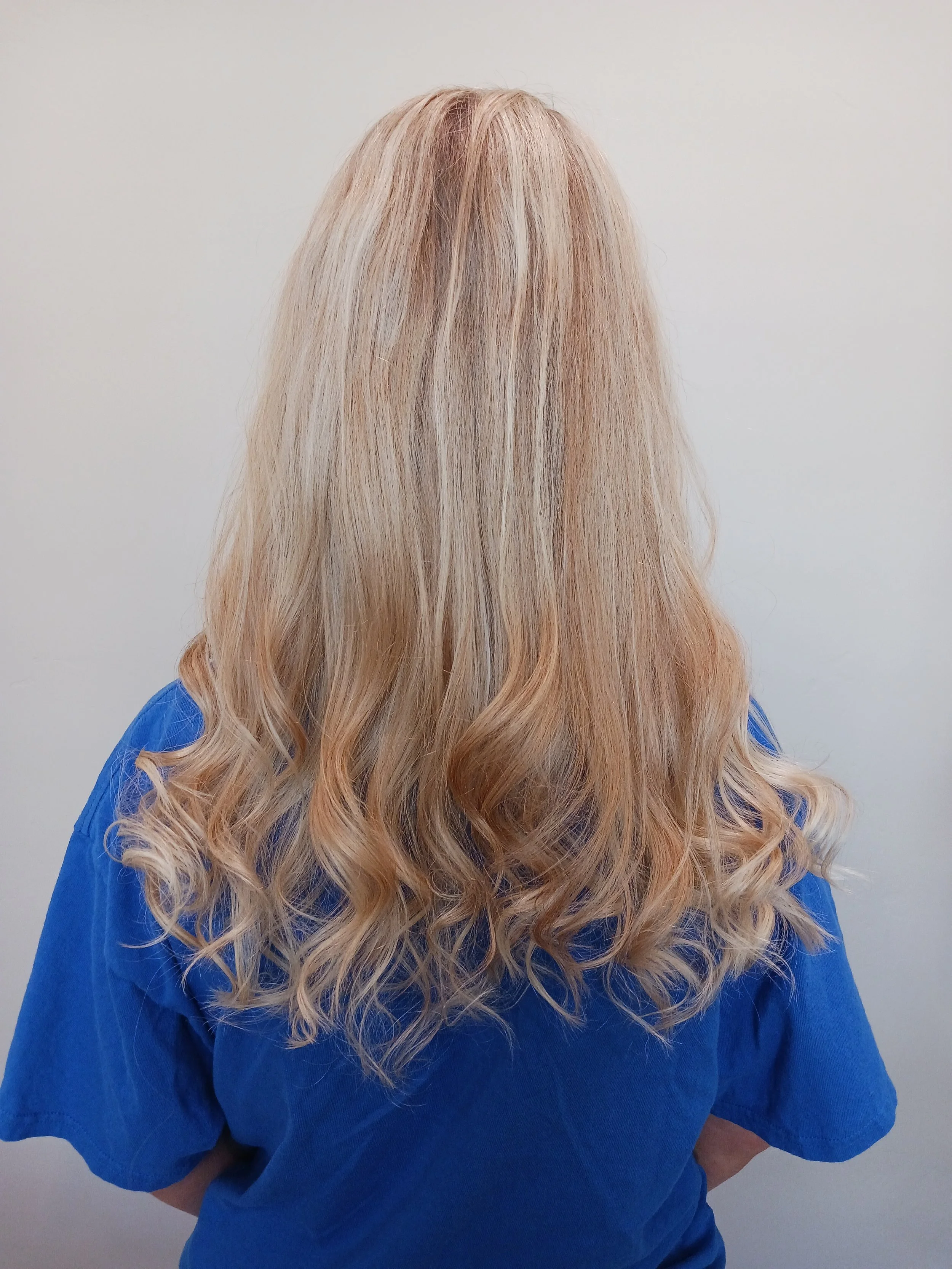 Back view of a woman with long, wavy blonde hair wearing a blue shirt, standing against a plain white wall.