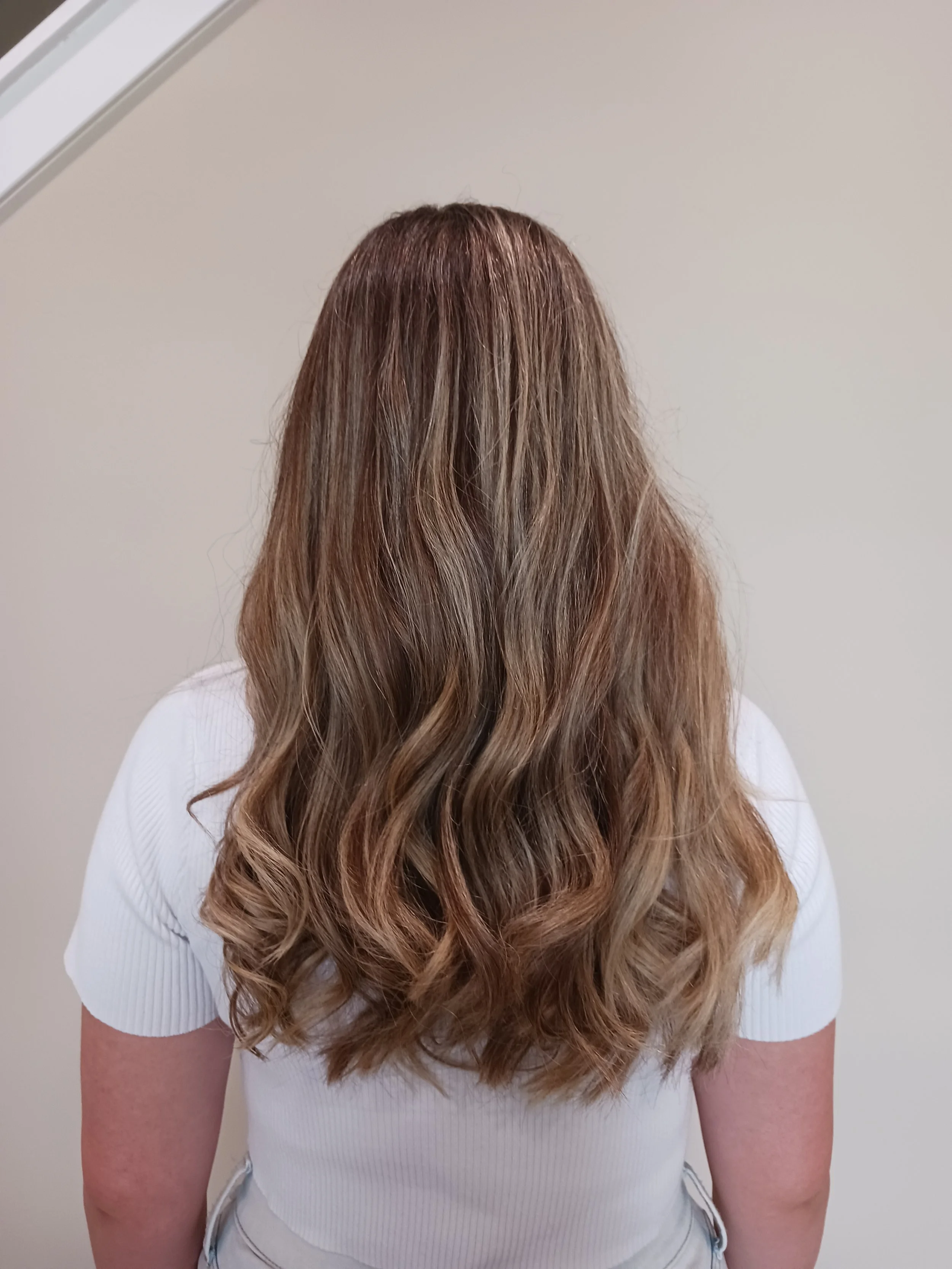 Back view of a woman with long, wavy, light brown hair standing against a plain wall.