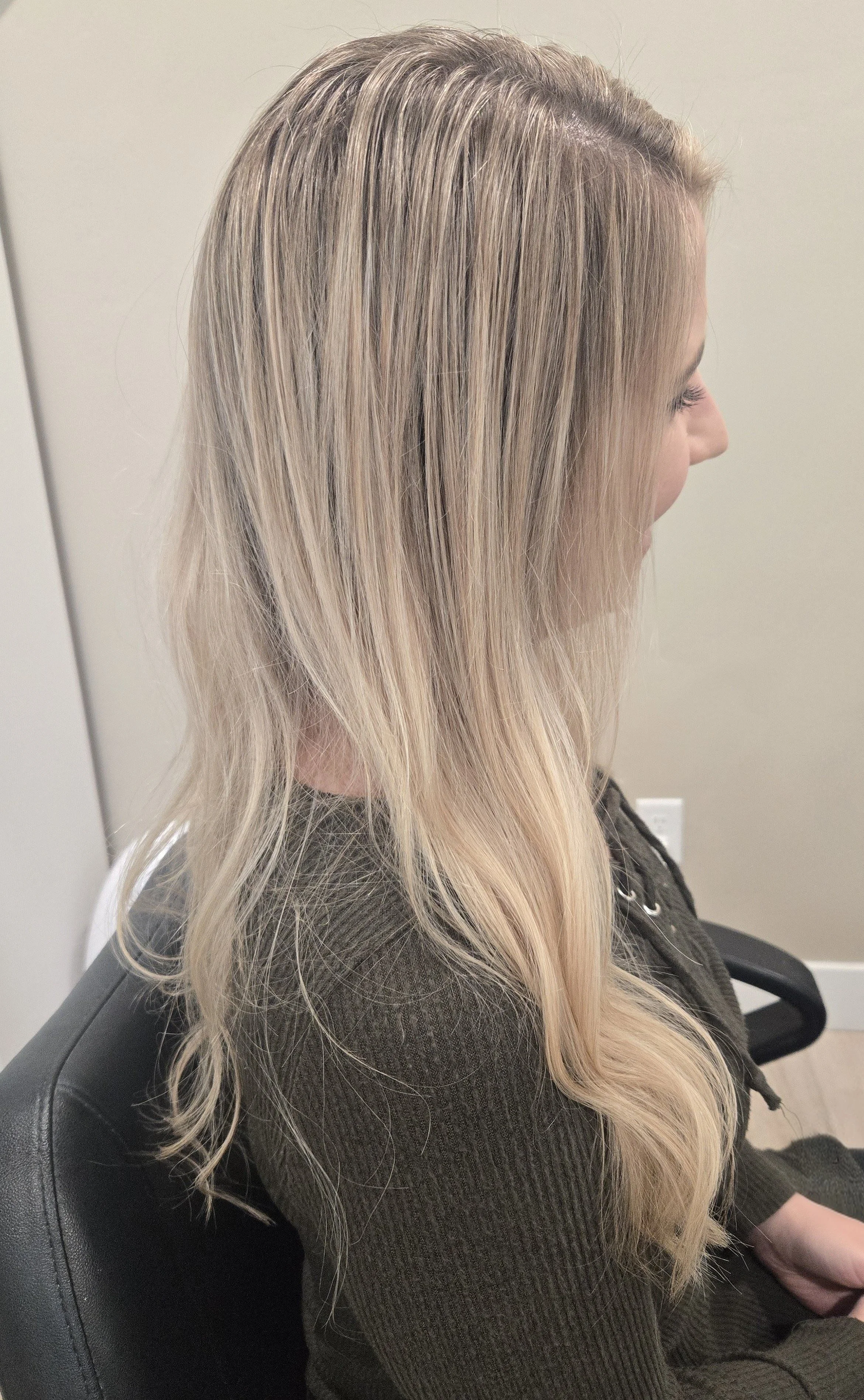 A side profile of a woman with long, wavy blonde hair sitting in a black chair in a room with beige walls.