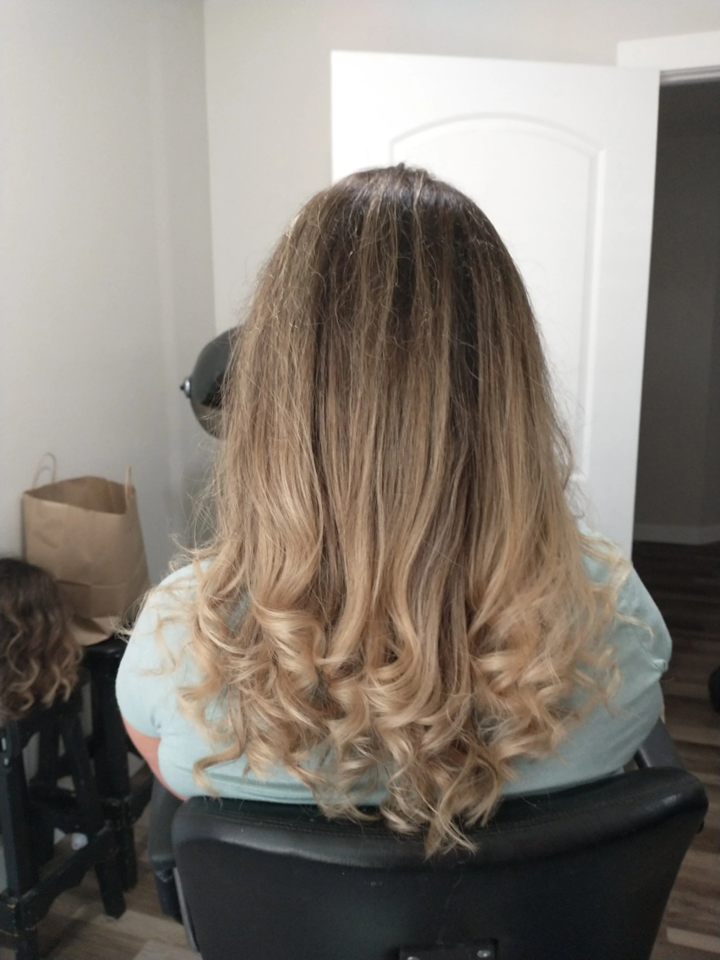 Back view of a woman with long, blonde curly hair sitting in a salon chair.